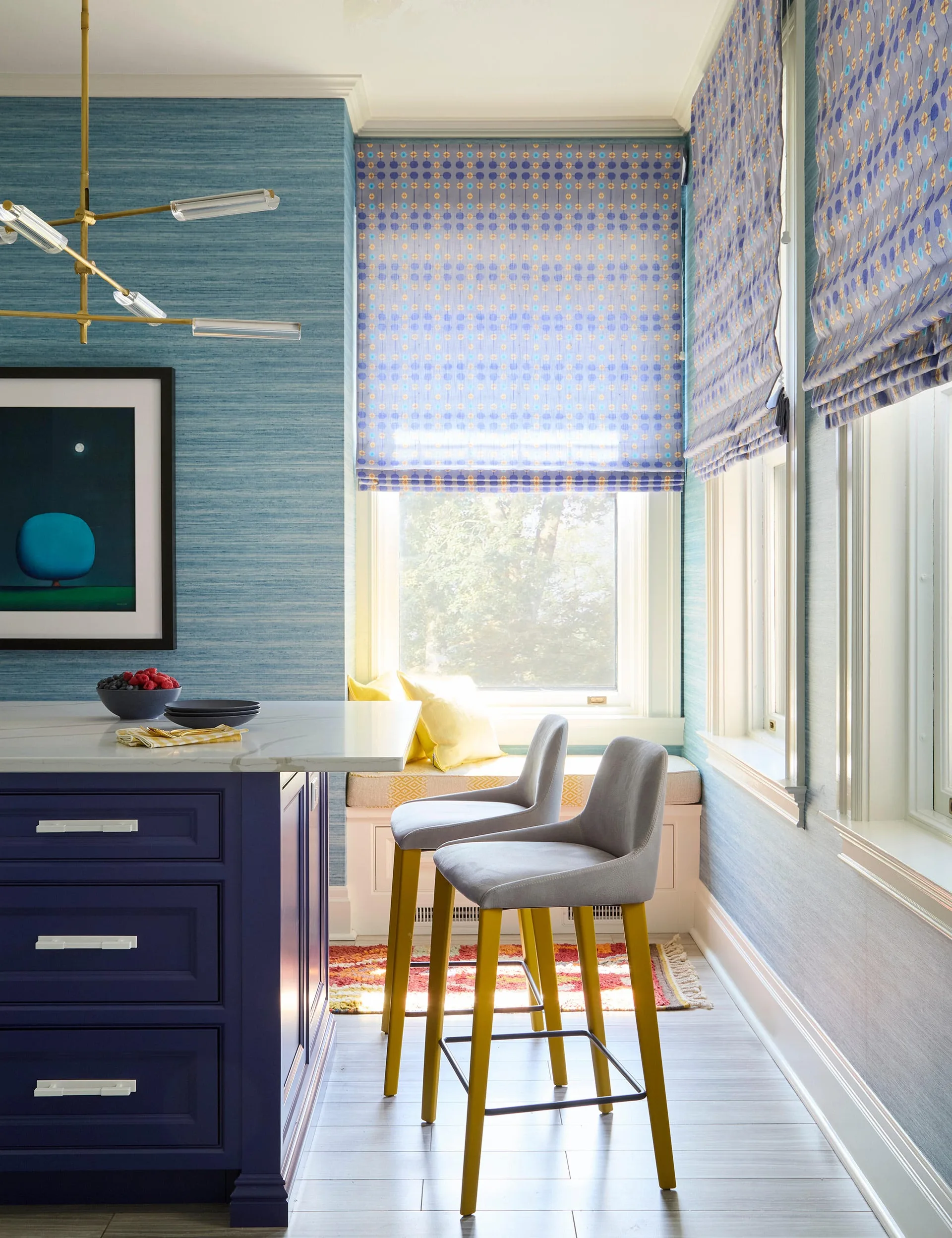 A blue kitchen island with a marble countertop and gray stools sits near a window seat with yellow pillows and patterned blue roman shades.