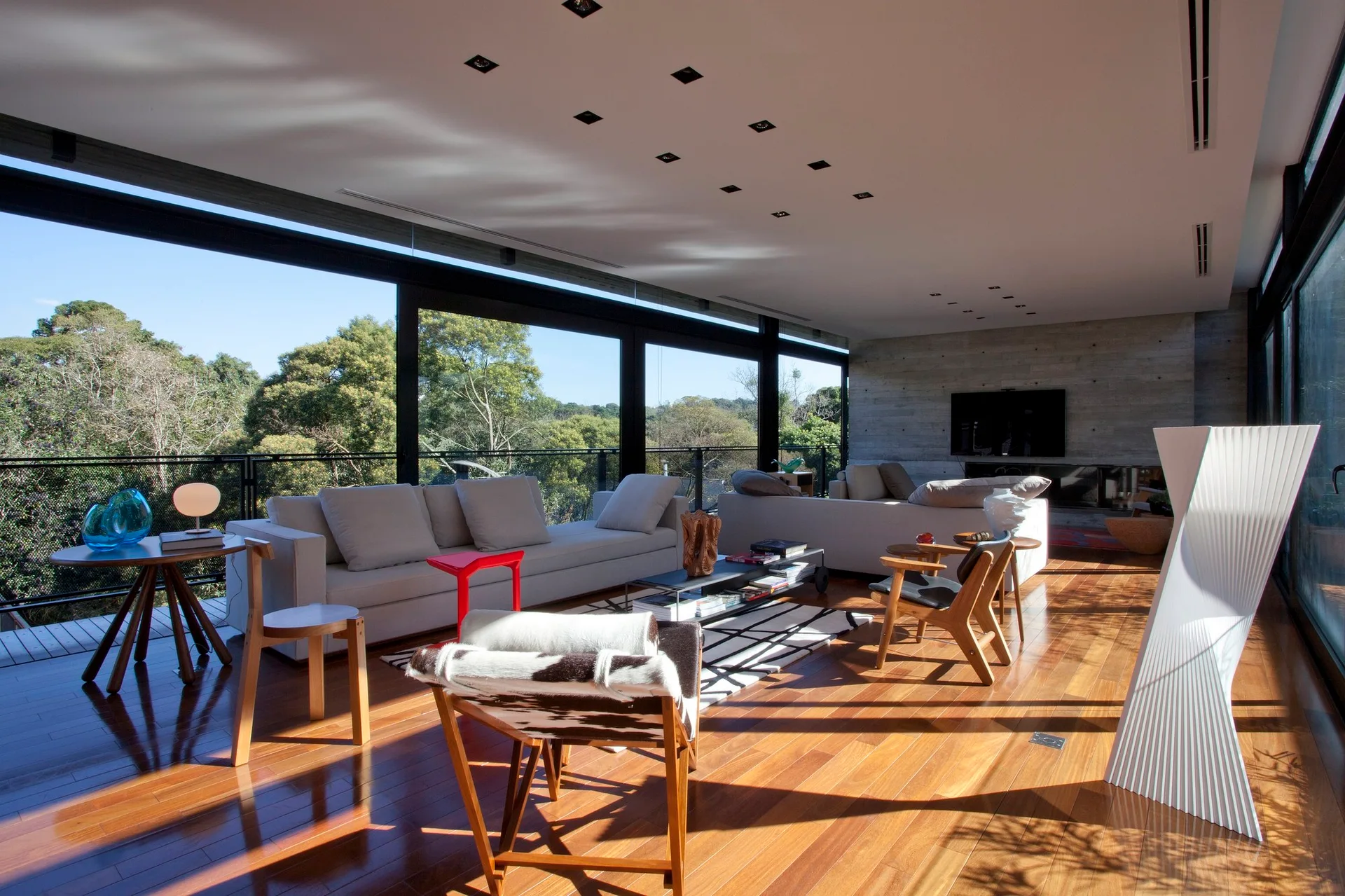 A spacious living room features off-white sofas, low-profile wooden chairs, and a polished wood floor next to large glass walls looking out at trees.