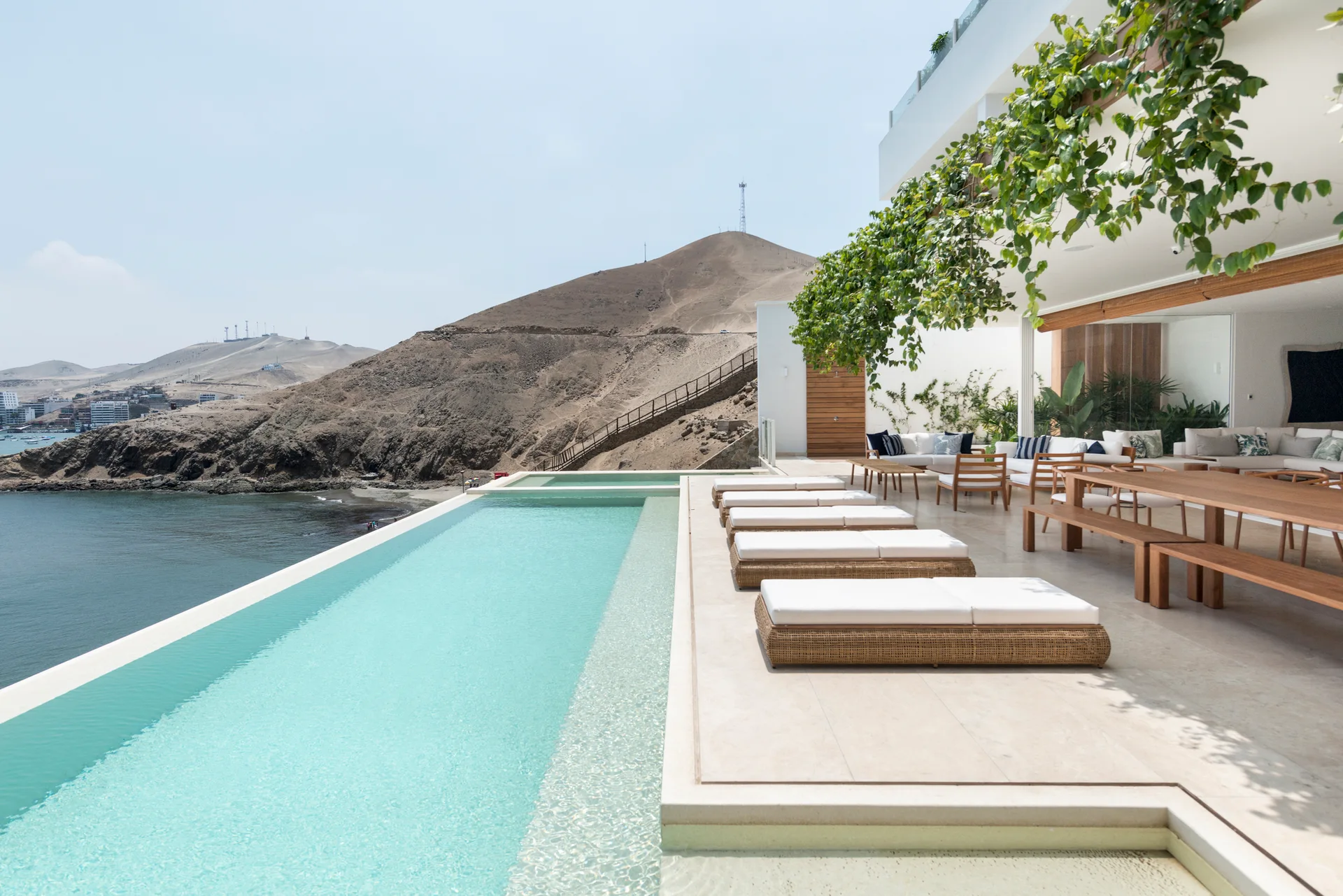 An outdoor infinity pool terrace overlooking the ocean features wicker lounge chairs, a wooden dining table, and a white modern building facade.