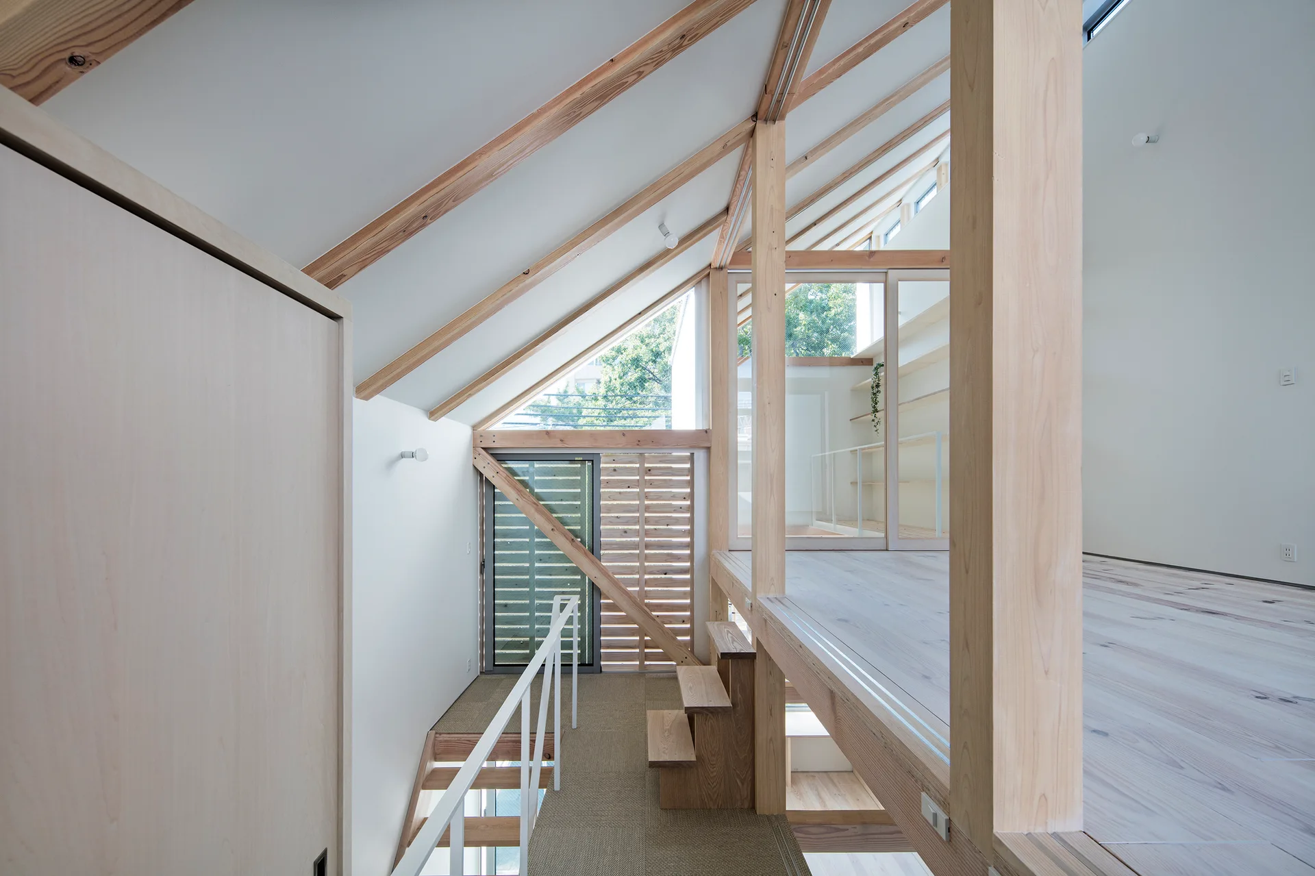A modern split-level loft features a wooden staircase with a white metal railing, exposed timber ceiling beams, and clean white walls.