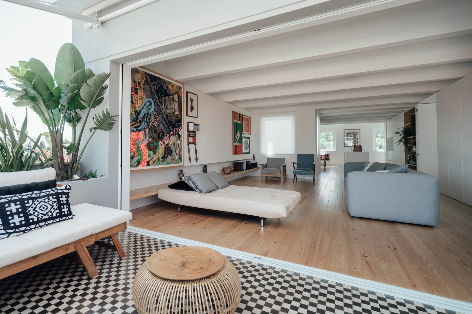 A modern living room with light wood floors and white ceiling beams opens onto a patio with checkerboard tile flooring.