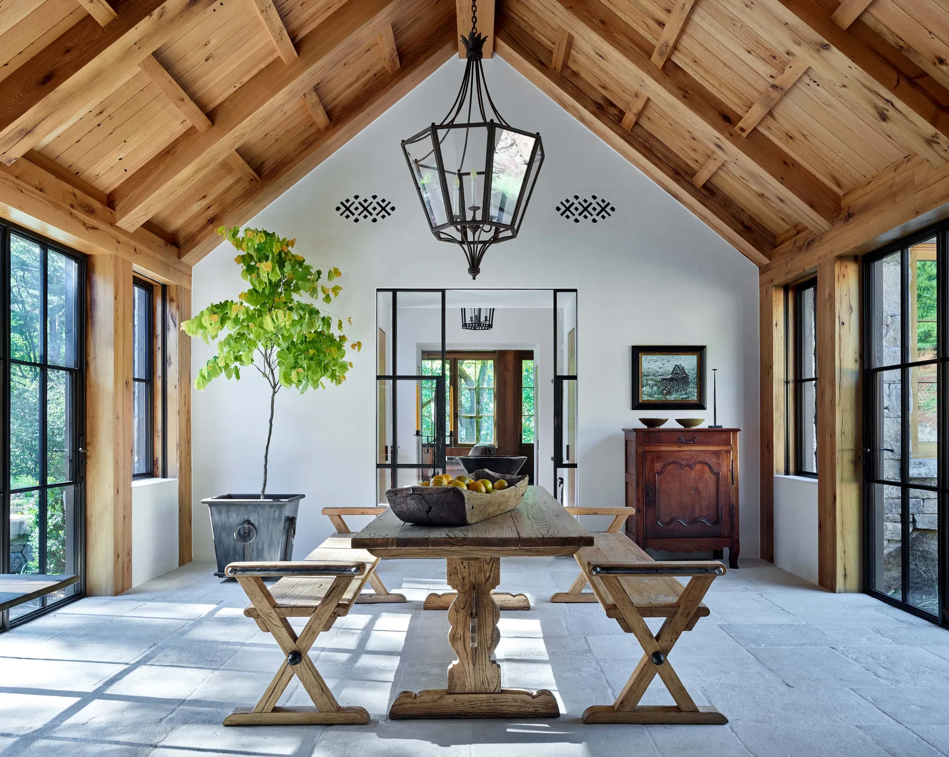 A rustic dining room featuring a vaulted wood ceiling, wooden table with benches, and large stone floor tiles under a black metal lantern.