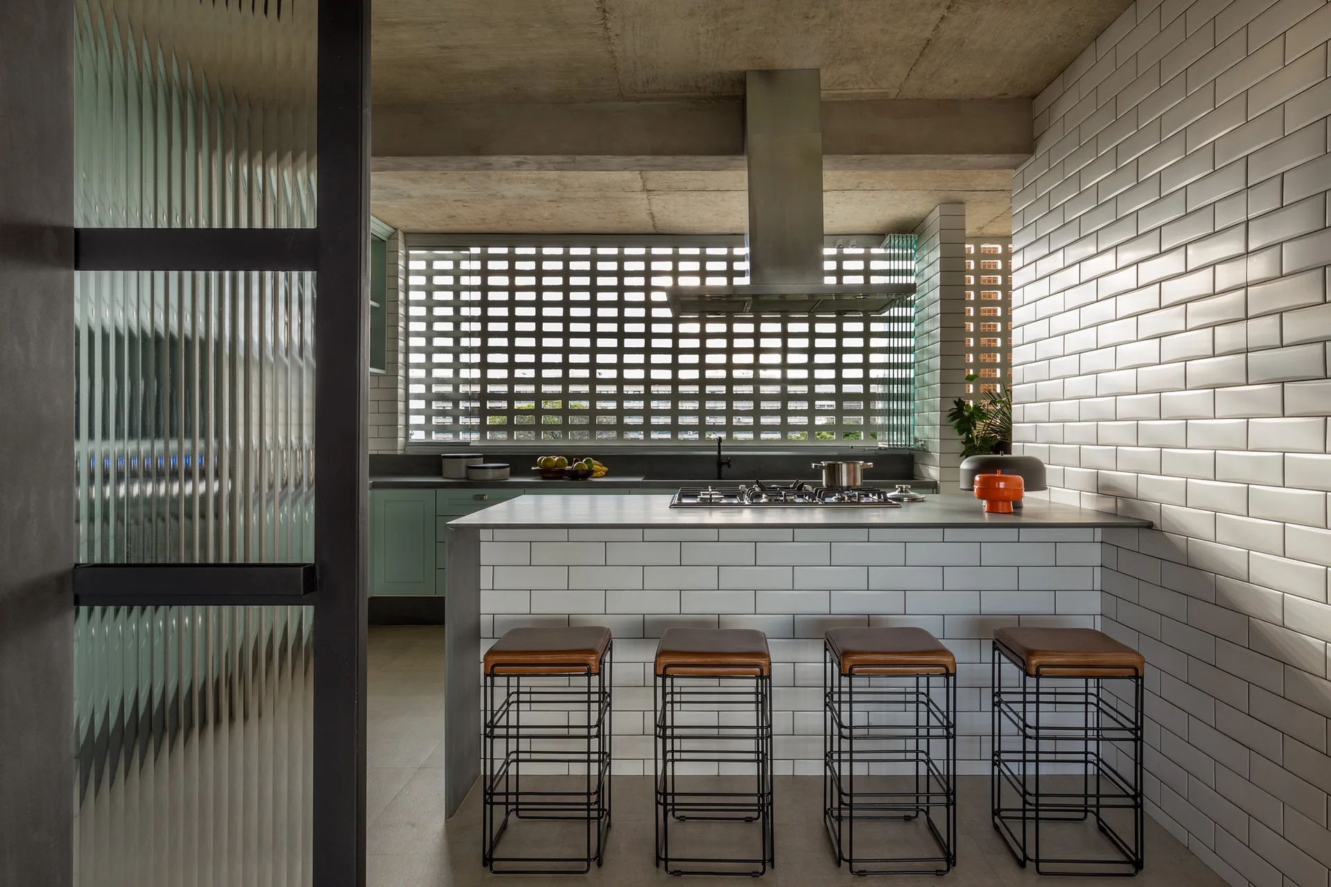 A modern kitchen featuring a subway tiled island with black bar stools, a concrete ceiling, and a breeze block wall.