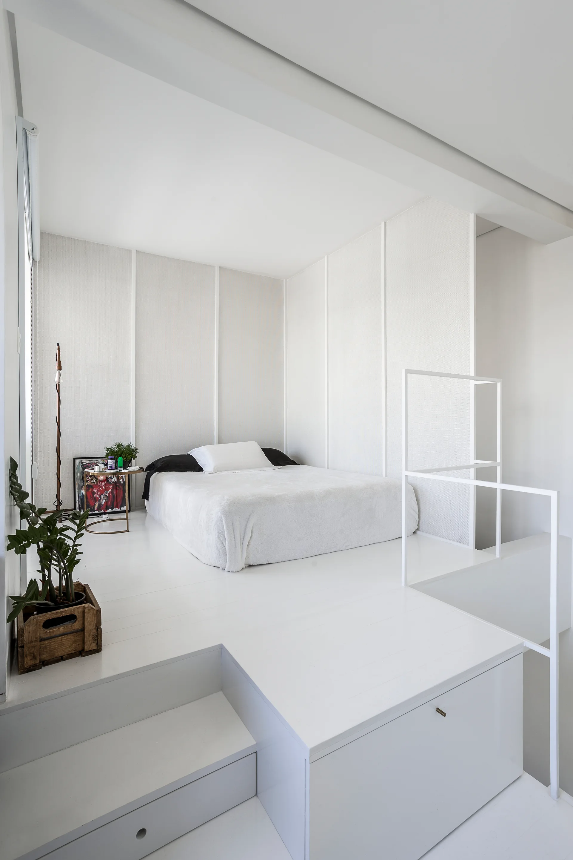 An all-white minimalist bedroom features a low-profile mattress on a raised platform with built-in drawers, vertical wall panels, and a metal railing.