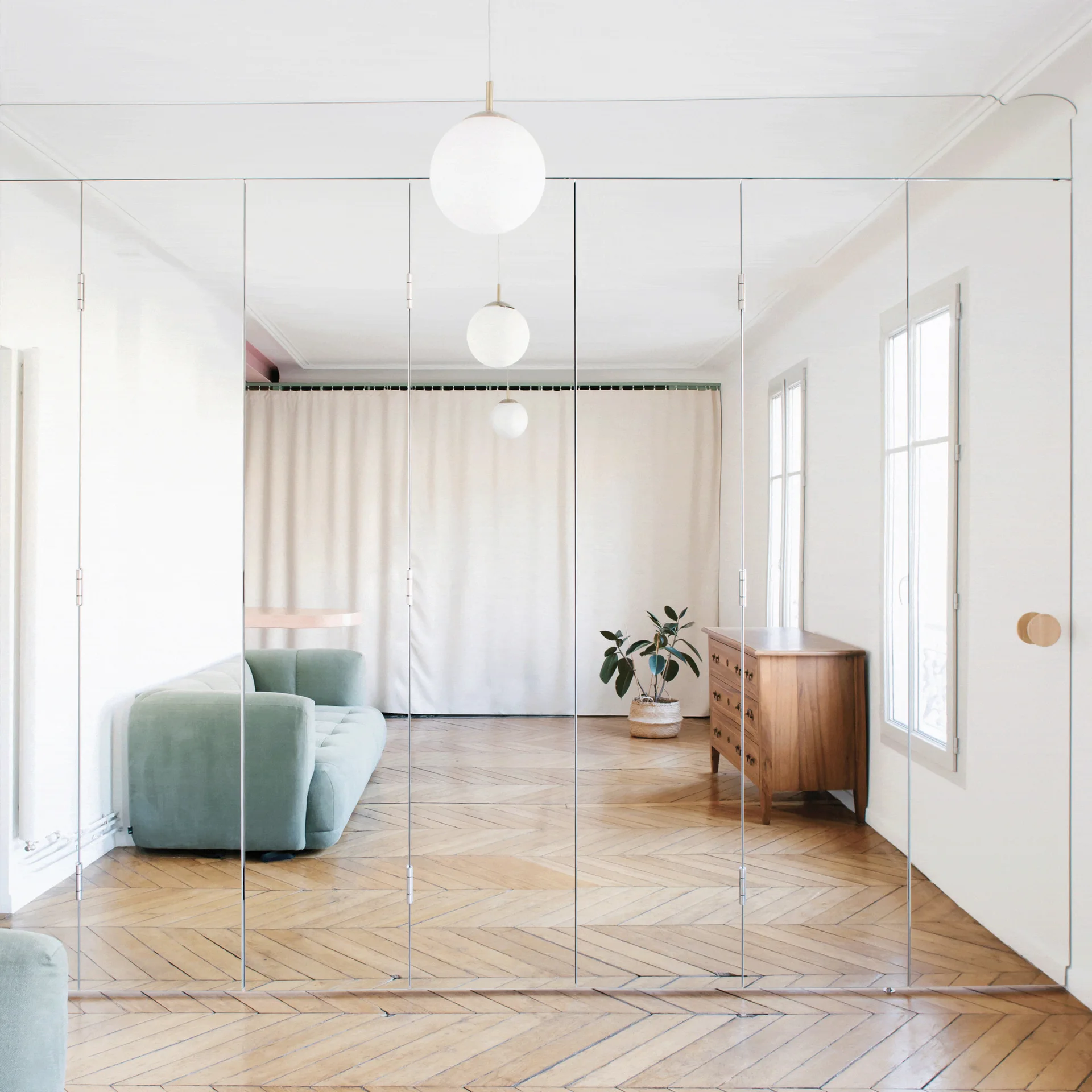 A bright minimalist living room featuring a green sofa and wood dresser reflected in a large floor-to-ceiling mirrored wall panel.