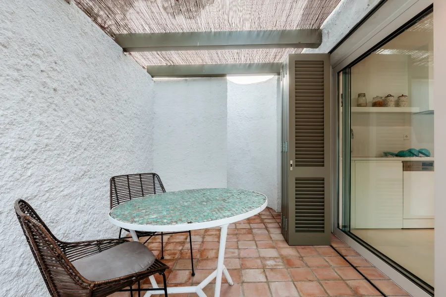 A small outdoor patio with terracotta tile flooring, rough white stucco walls, a reed-covered pergola, and a green mosaic bistro table.
