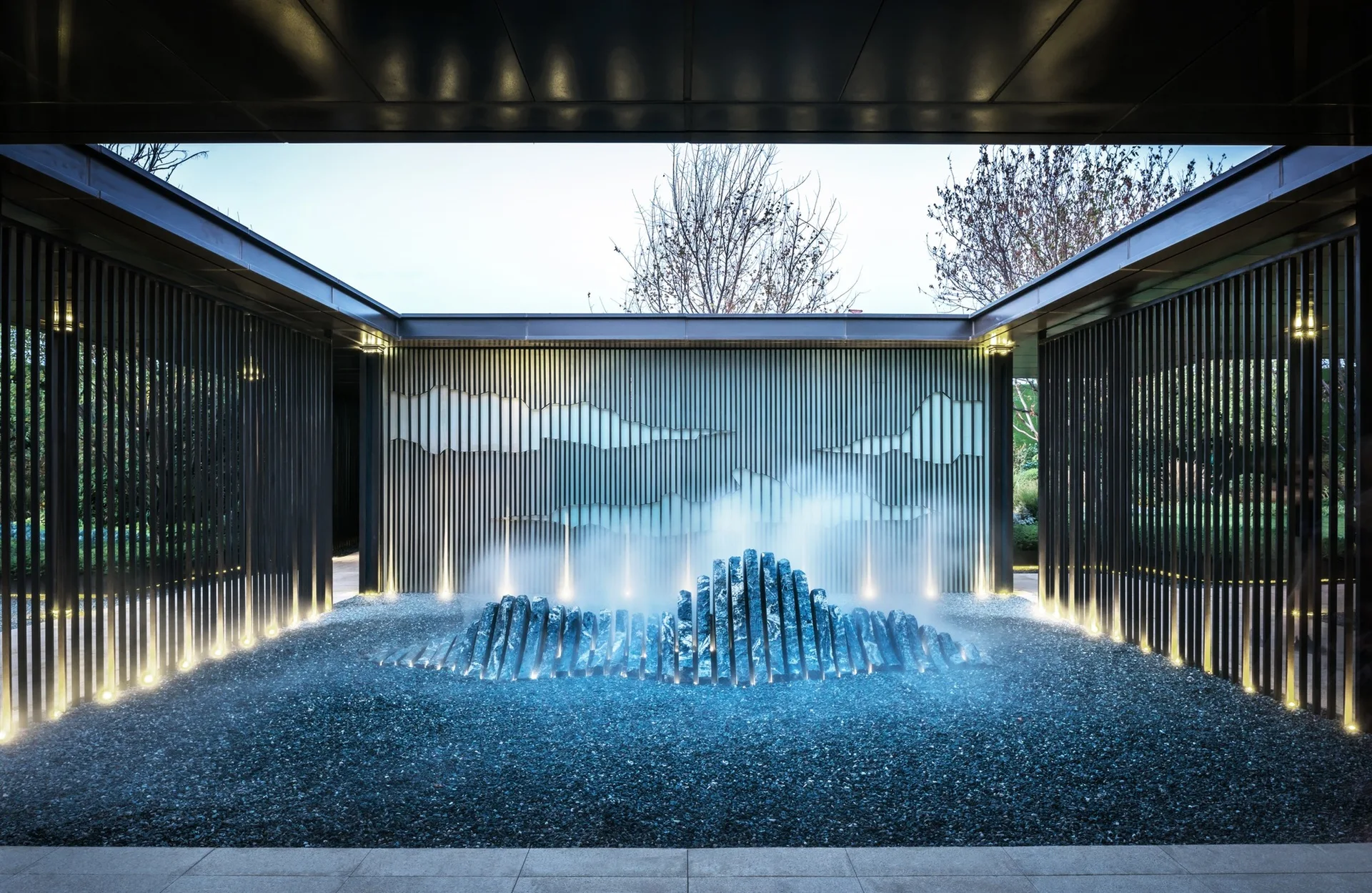 A modern courtyard features a central misting stone sculpture on a gravel bed framed by dark slatted walls with integrated uplighting.