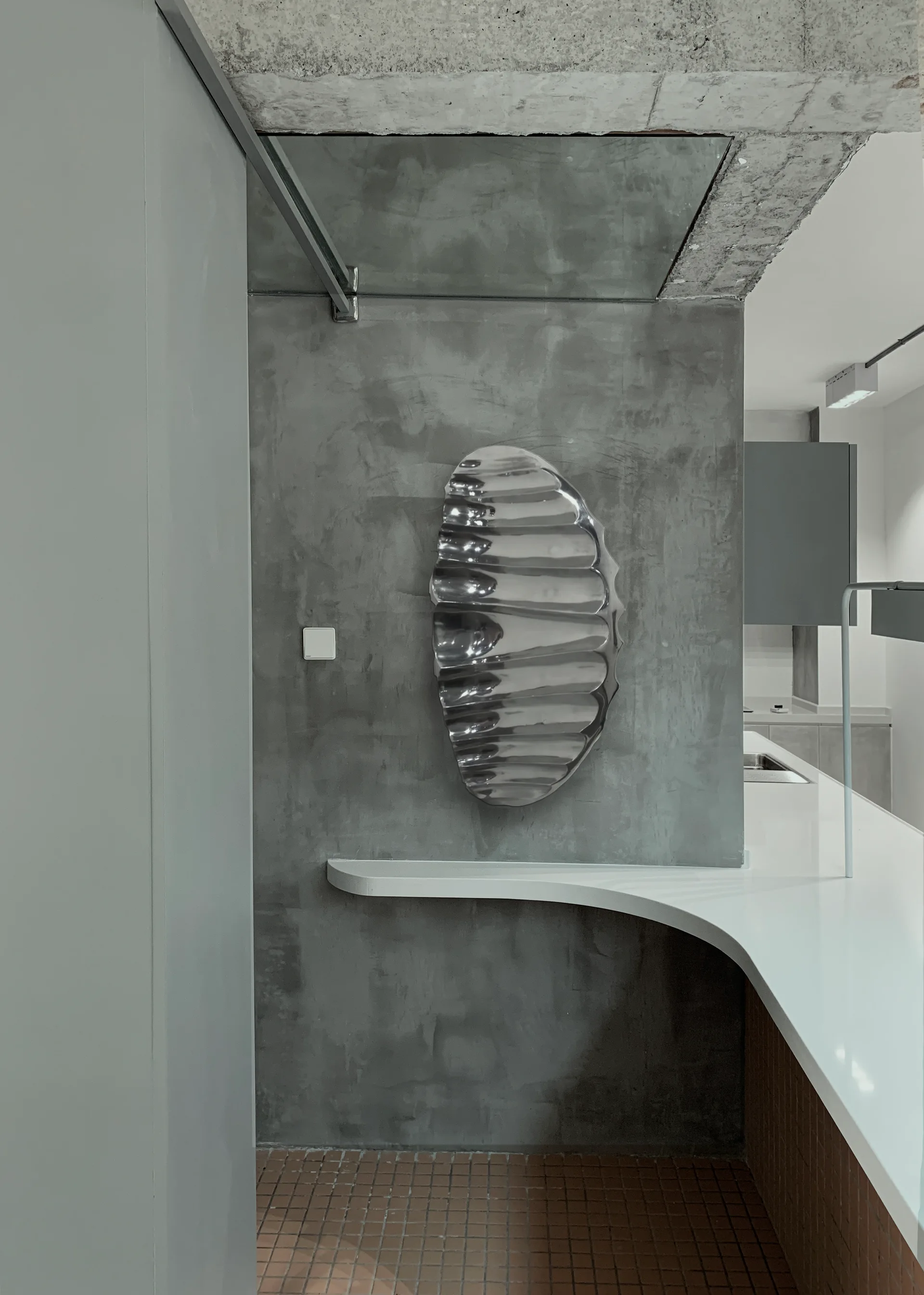 A modern kitchen corner featuring a textured gray concrete wall with a wavy silver sculpture above a curved white countertop and terracotta tiled floor.