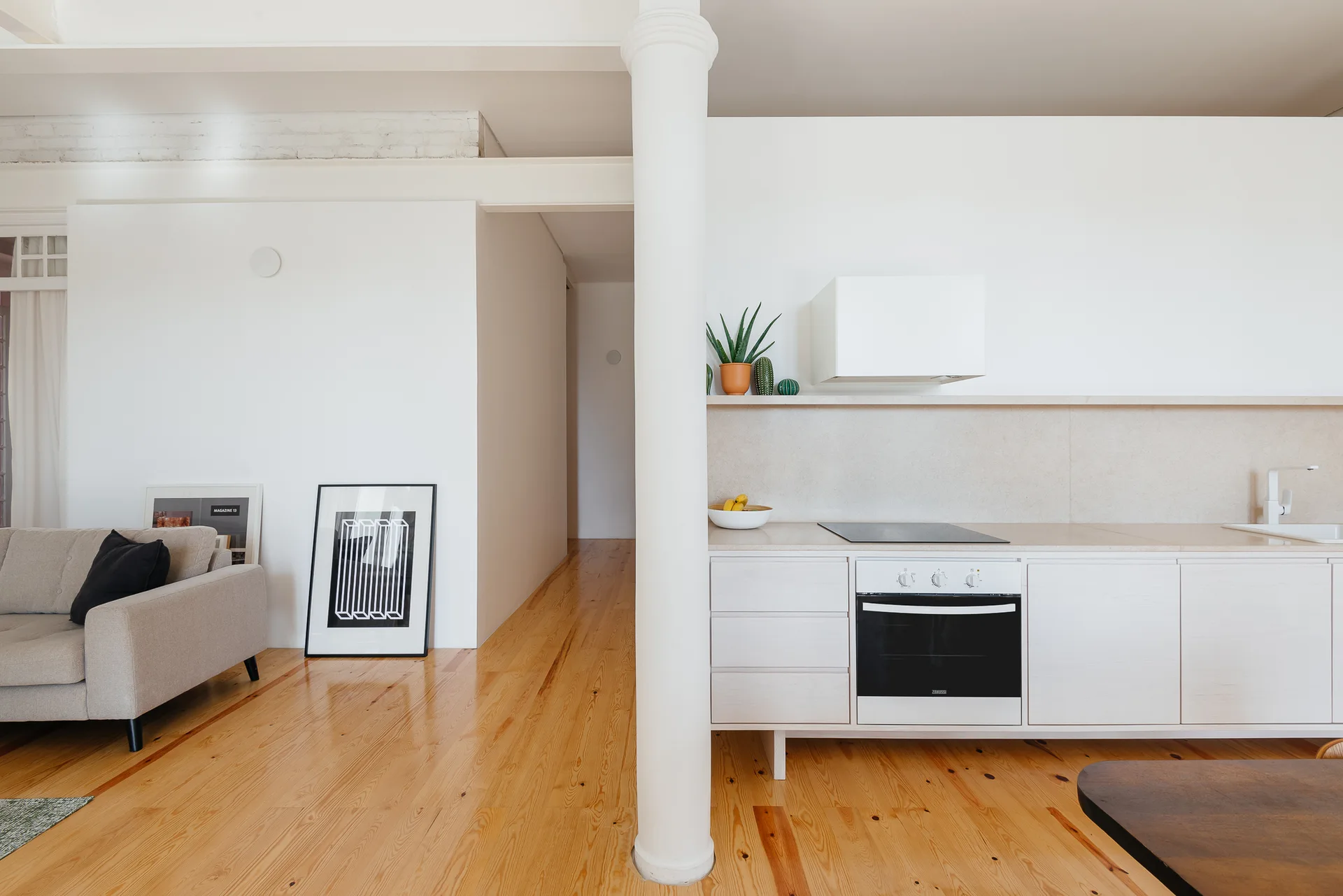 A minimalist open-concept kitchen features white flat-front cabinets, a central white column, and pine wood floors next to a light grey sofa.