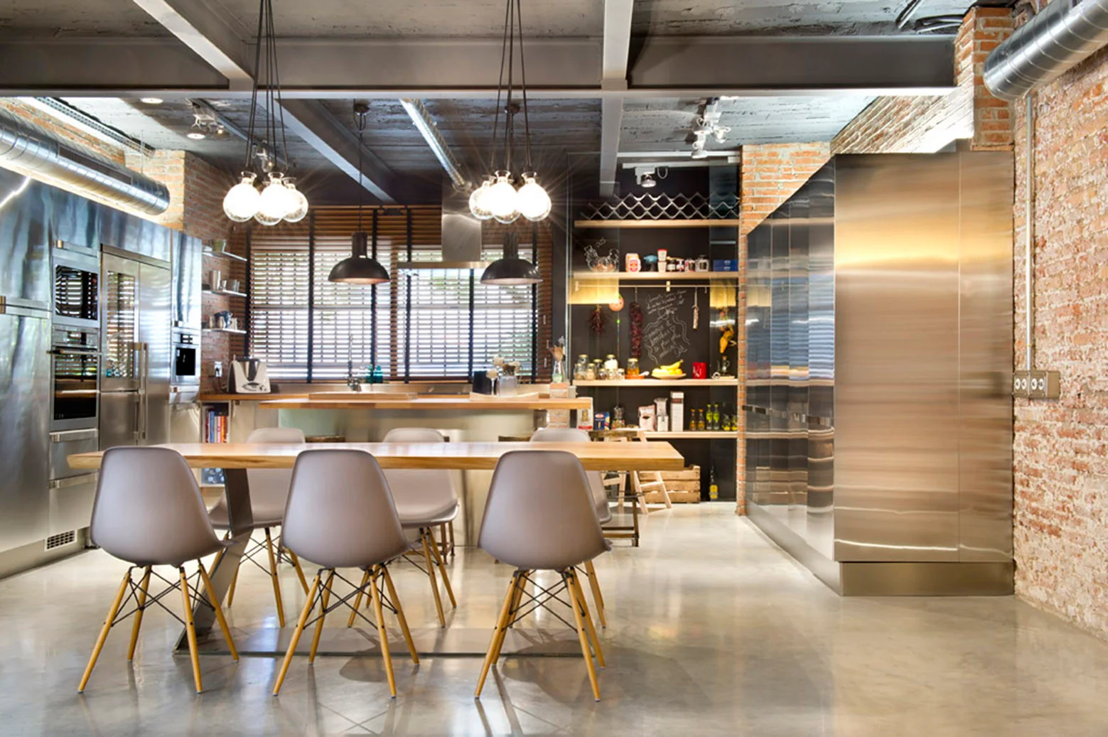 An industrial kitchen with stainless steel cabinets, a red brick wall, and a wooden dining table with grey chairs on a polished concrete floor.