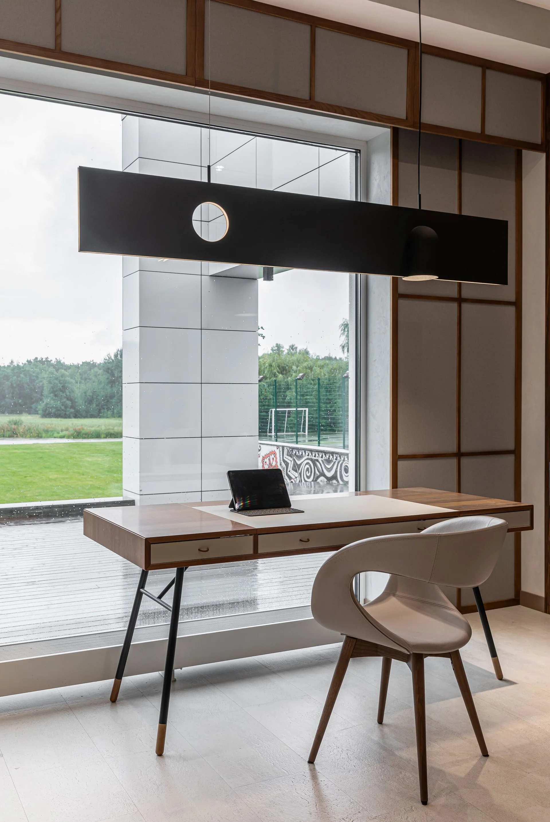 A modern home office features a wood desk, a curved beige chair, and a large black rectangular pendant light in front of a floor-to-ceiling window.