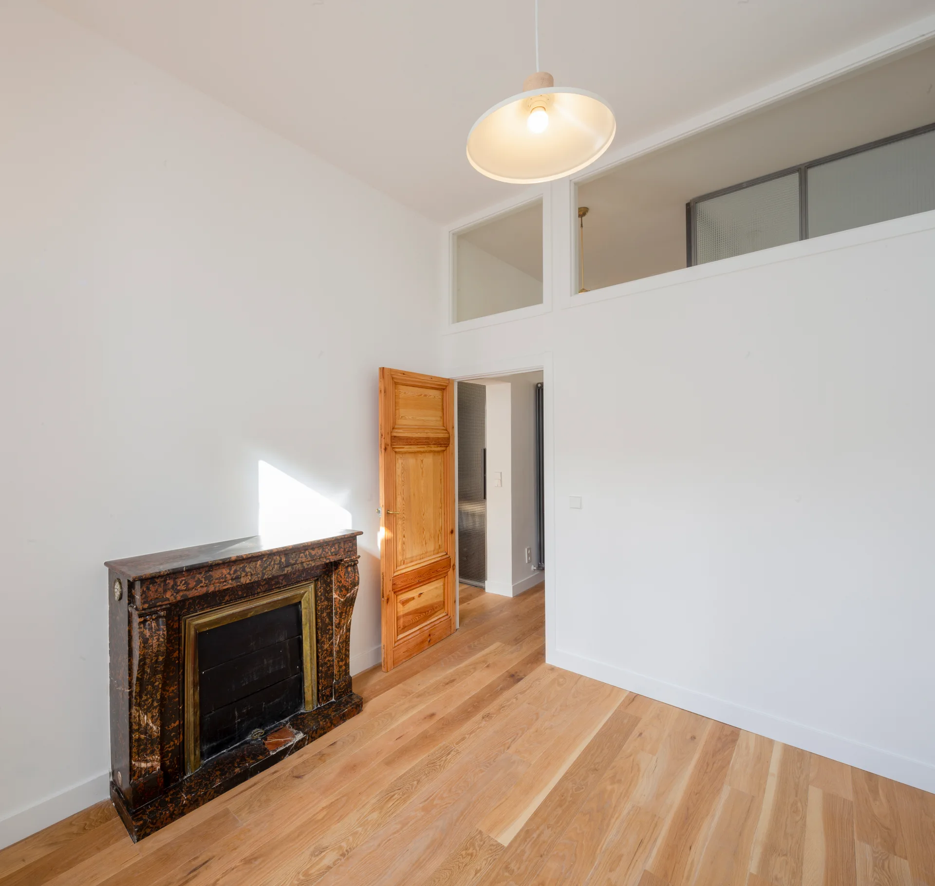 A minimalist white room features light oak hardwood flooring, an ornate dark marble fireplace, and a natural wood door below a glass transom window.