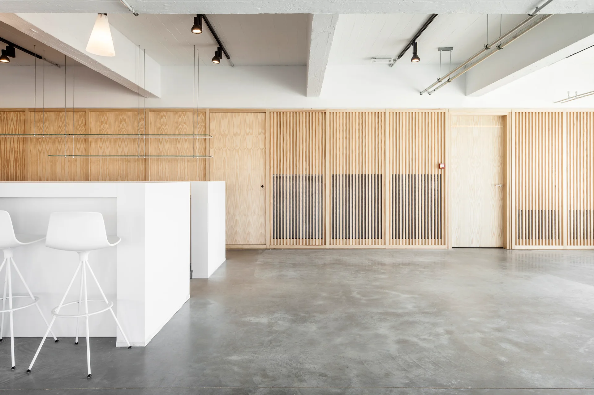 A minimalist white kitchen island with matching bar stools stands on a grey concrete floor in front of a light wood slatted wall.