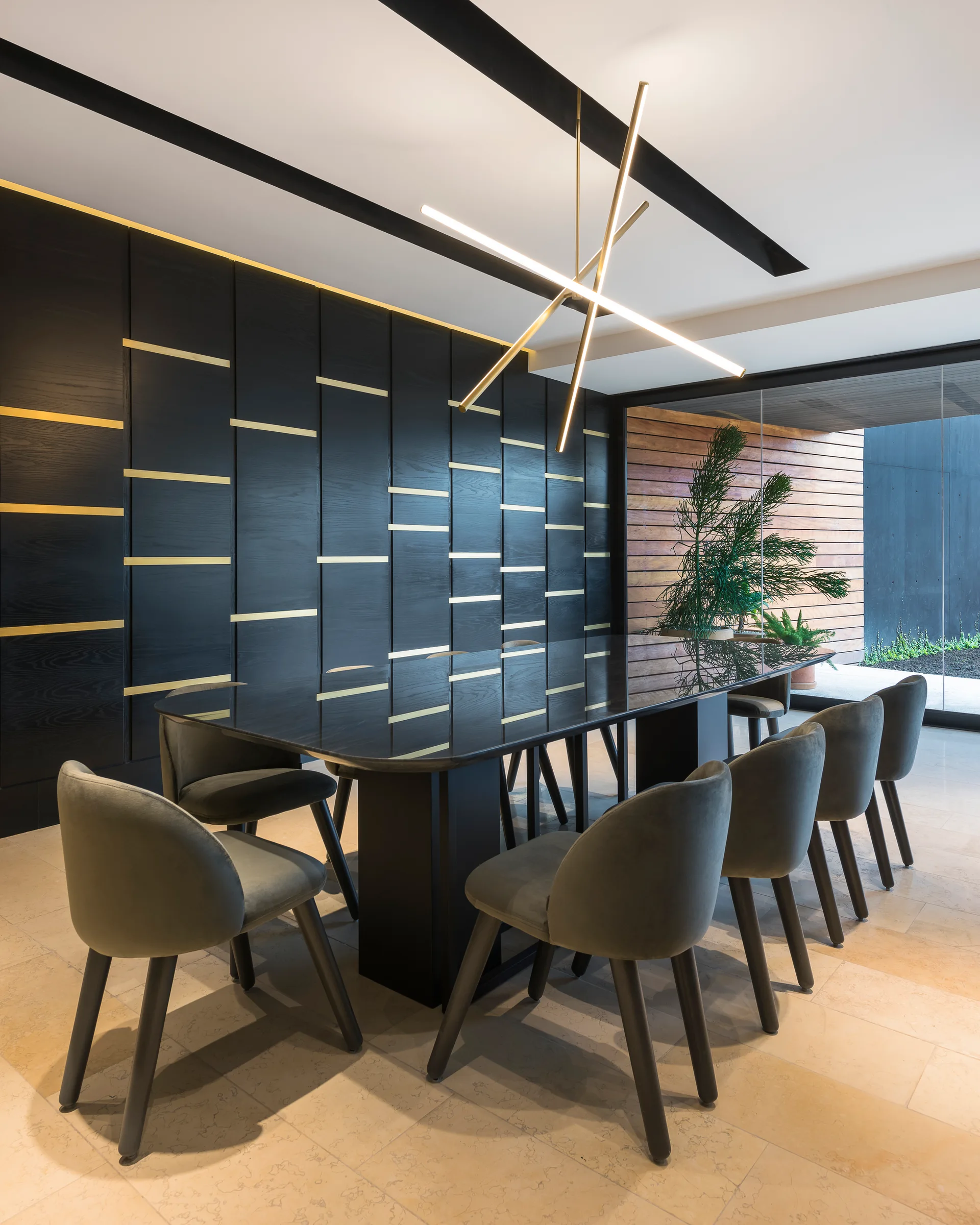 A modern dining room features a glossy black table surrounded by gray upholstered chairs in front of a dark wood paneled wall with gold linear accents.