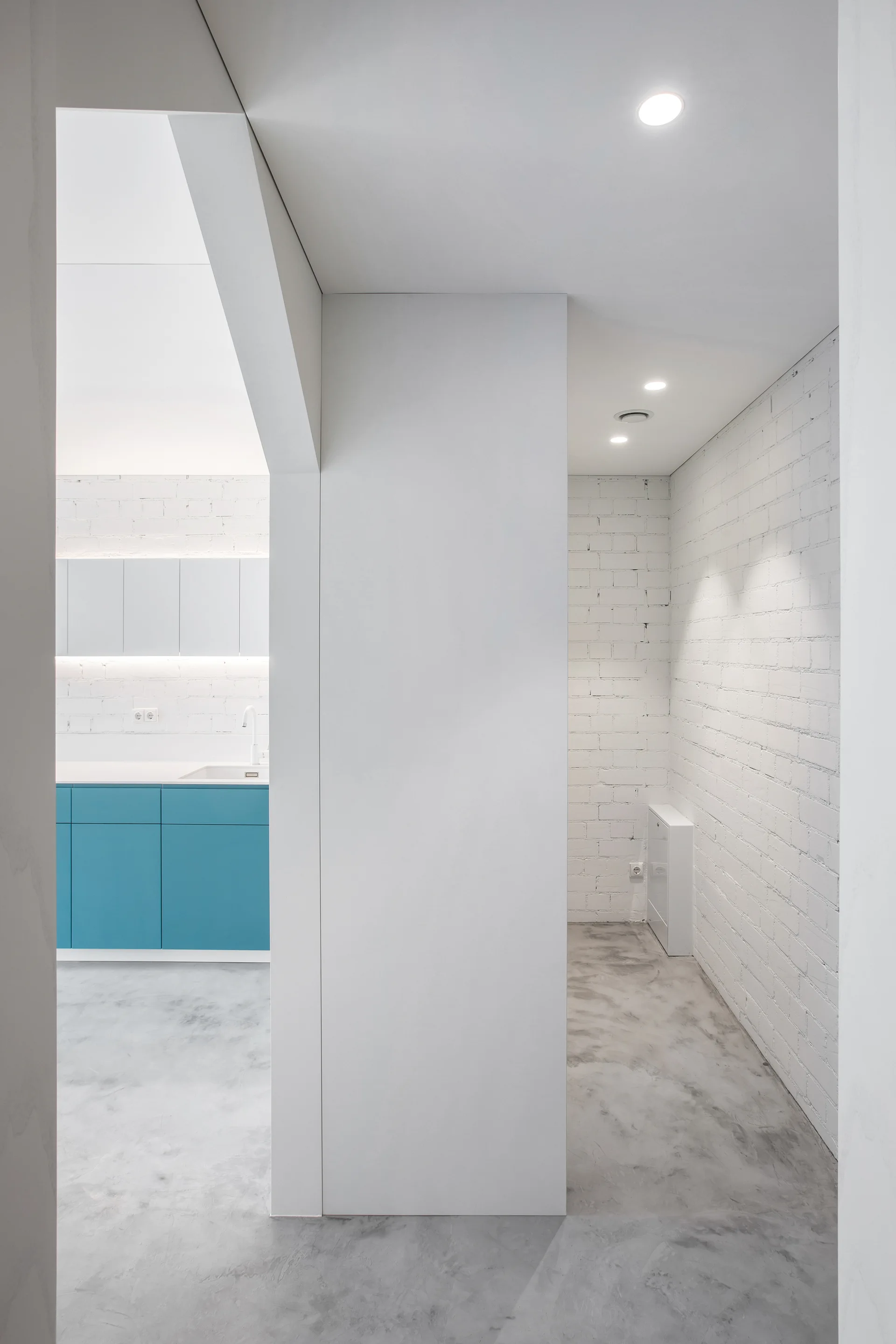 A view through a minimalist white doorway of a kitchen featuring blue lower cabinets, white painted brick walls, and a polished concrete floor.