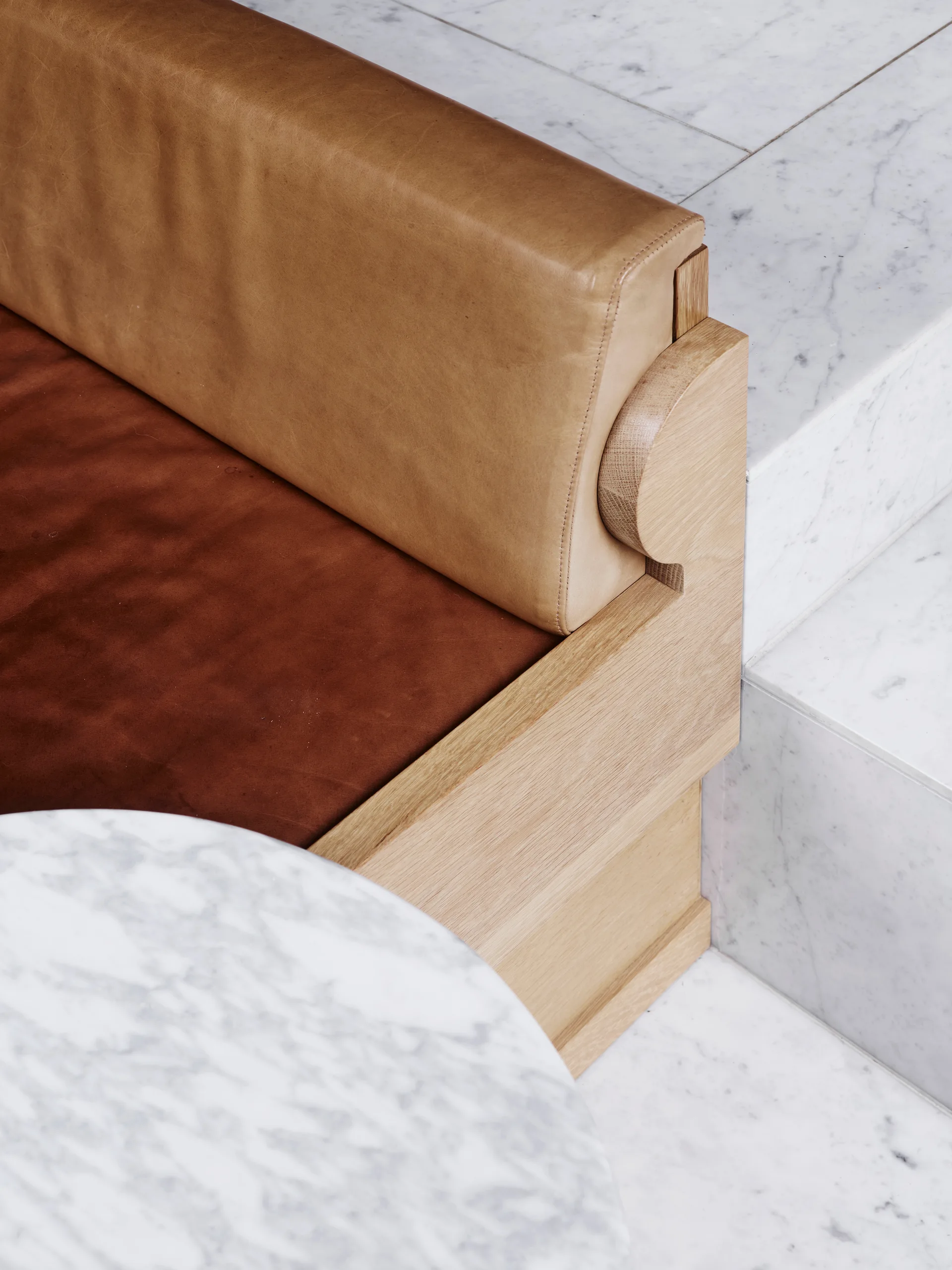A close-up of a custom wood sofa with tan and brown leather cushions next to a round marble table on a white marble floor.