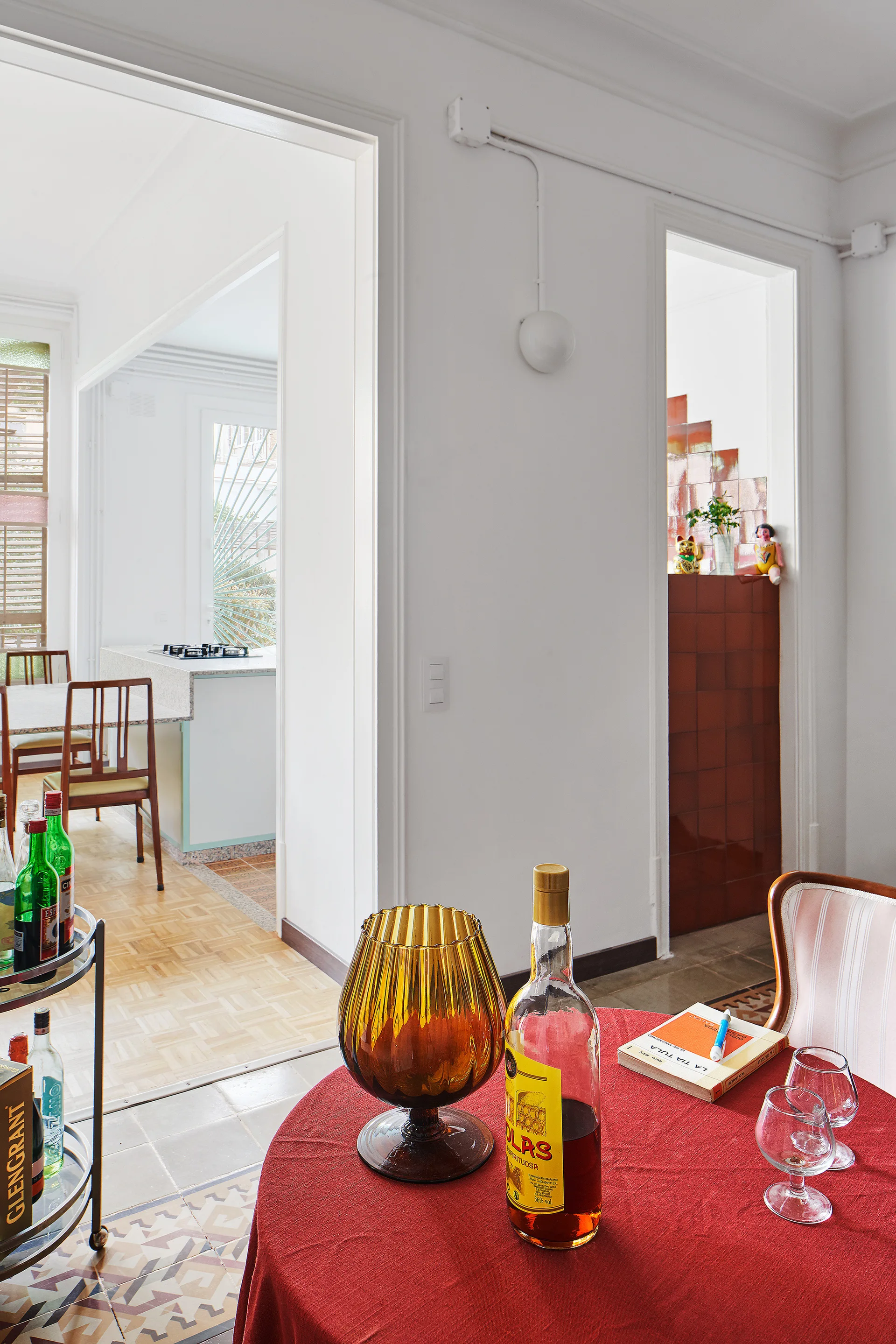 A dining table with a red tablecloth, amber glass vase, and drinks sits on patterned tile floors near doorways leading to a kitchen.