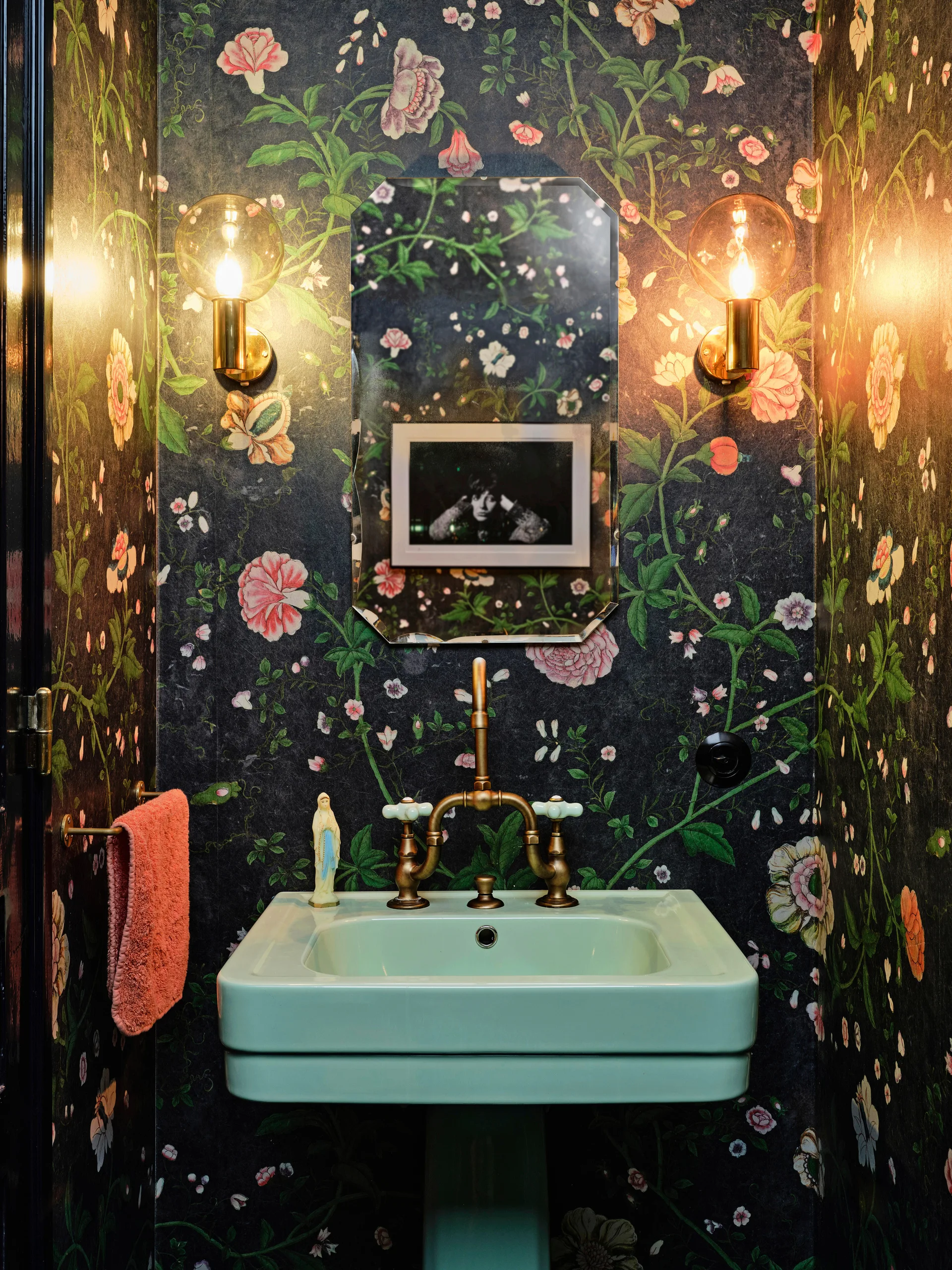 A small powder room with dark floral wallpaper, a mint green pedestal sink with brass hardware, and round brass sconces flanking an octagonal mirror.