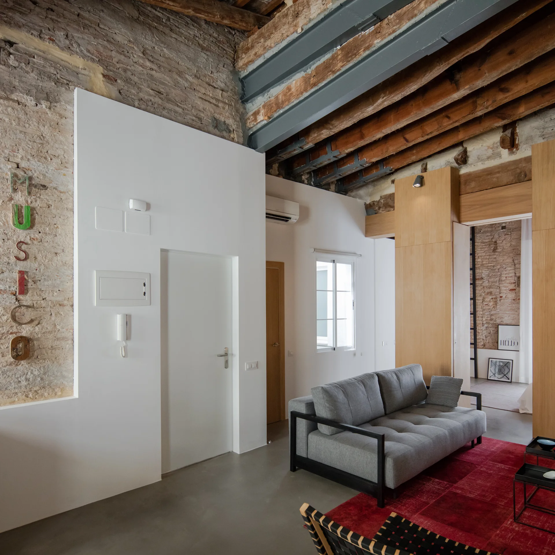 An industrial living room features a grey sofa on a red rug beneath an exposed timber ceiling, alongside white walls and exposed brick.