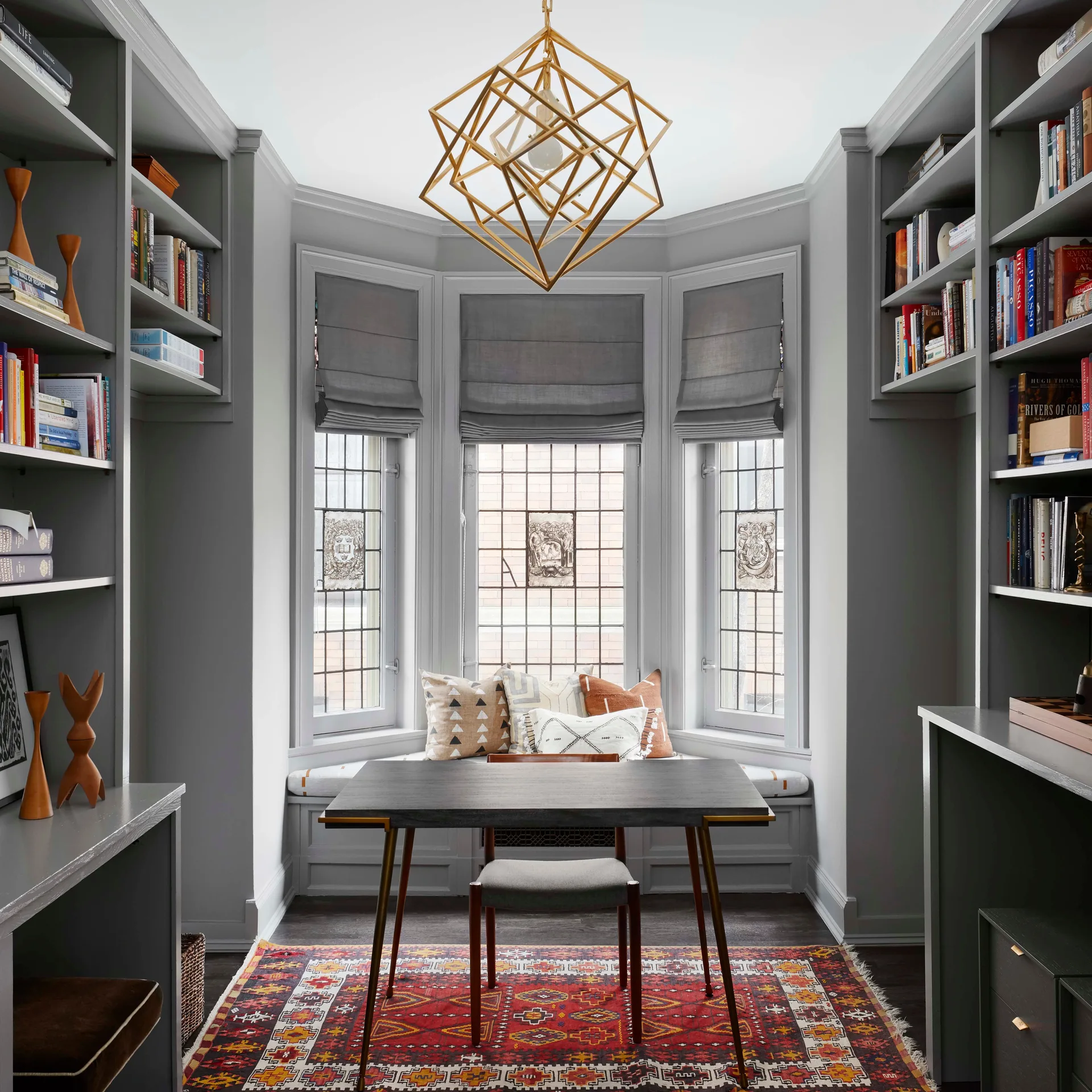 A symmetrical gray home office featuring floor-to-ceiling bookshelves, a bay window with a cushioned seat, a central desk, and a red patterned area rug.