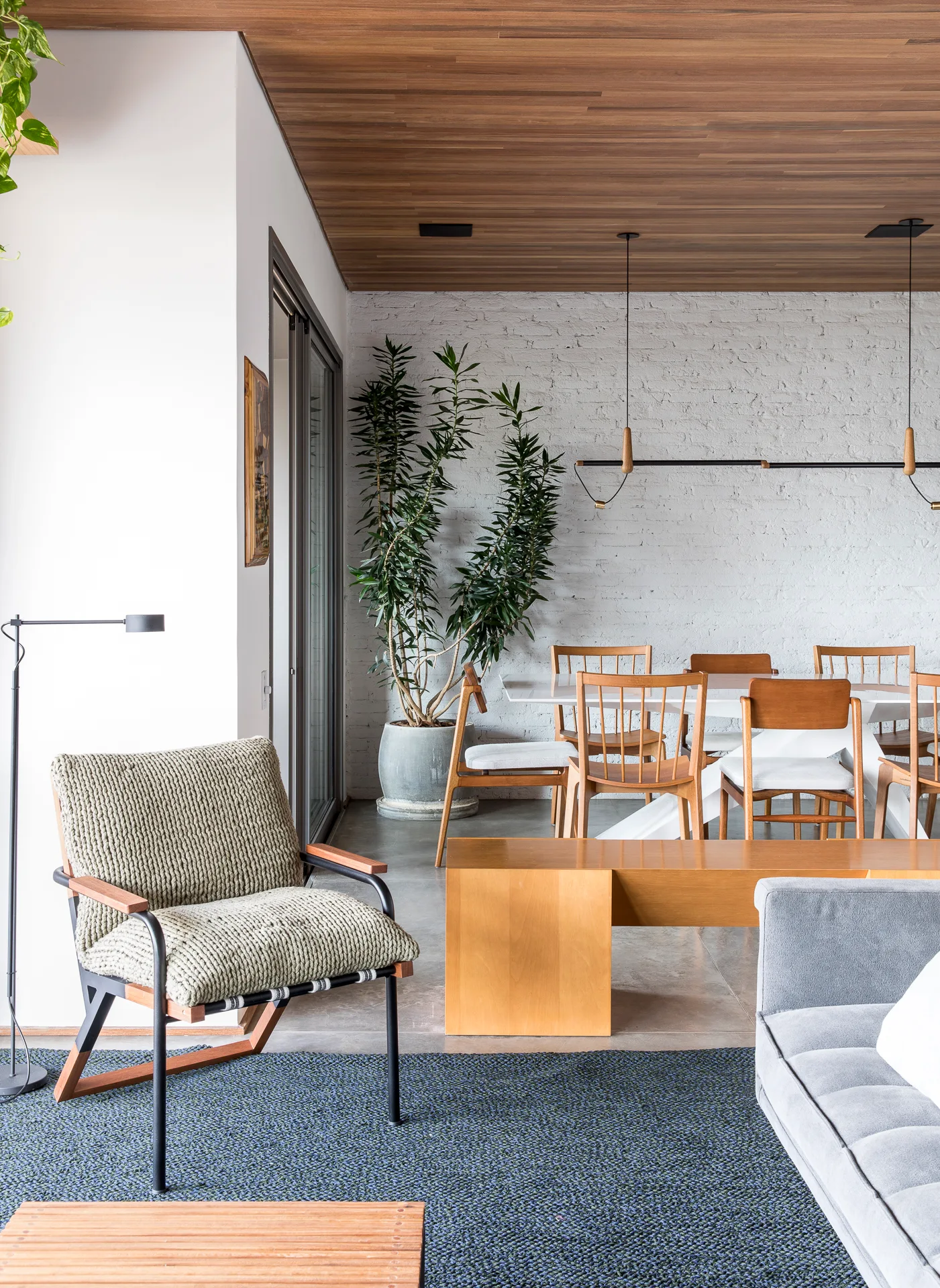 An open living and dining area featuring a white brick wall, warm wood plank ceiling, and mid-century modern wooden dining chairs.