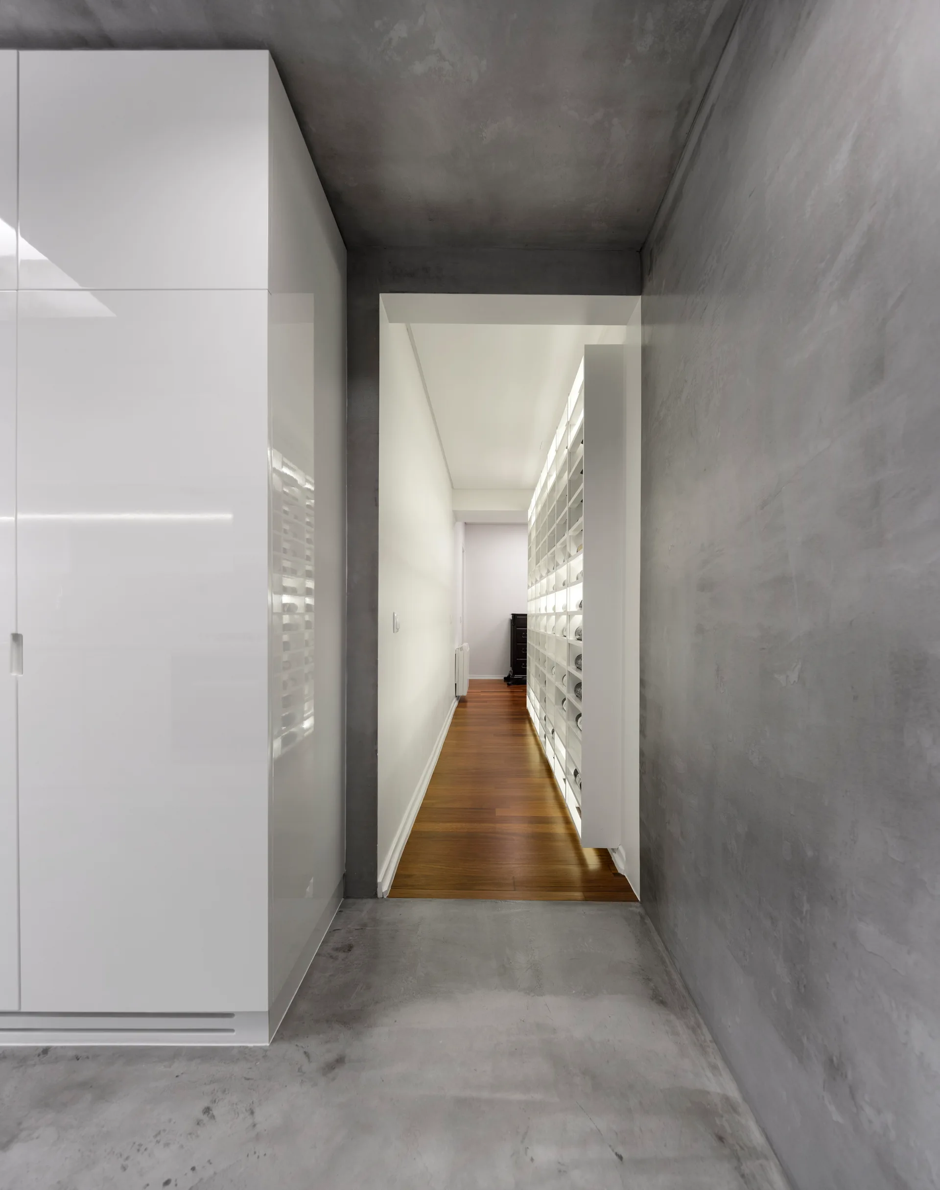 A modern hallway with concrete walls and floor leads through an open doorway to a corridor with warm wood flooring and illuminated shelving.