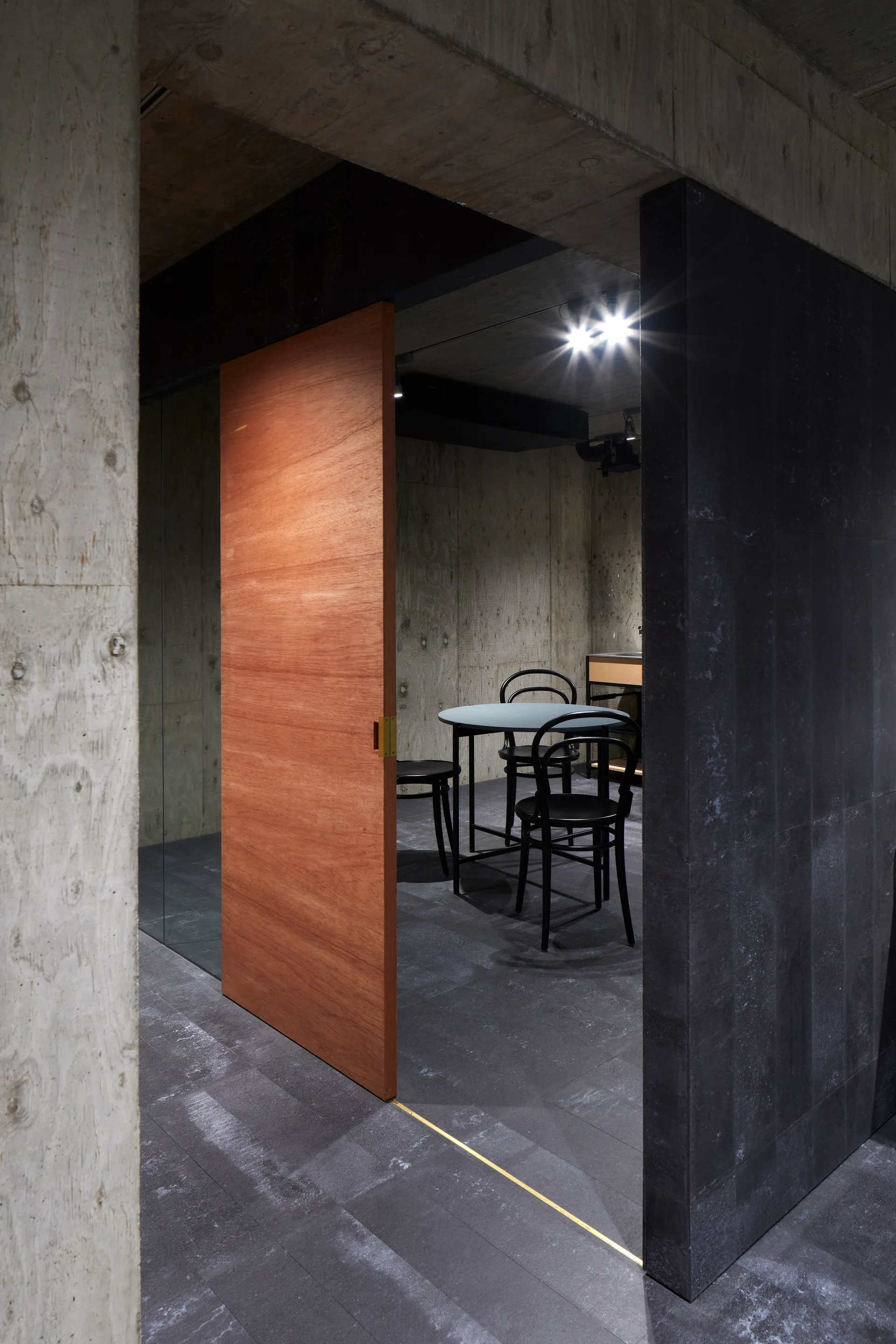 A modern dining room with raw concrete walls, a large wooden pivot door, and black chairs arranged around a round grey table.