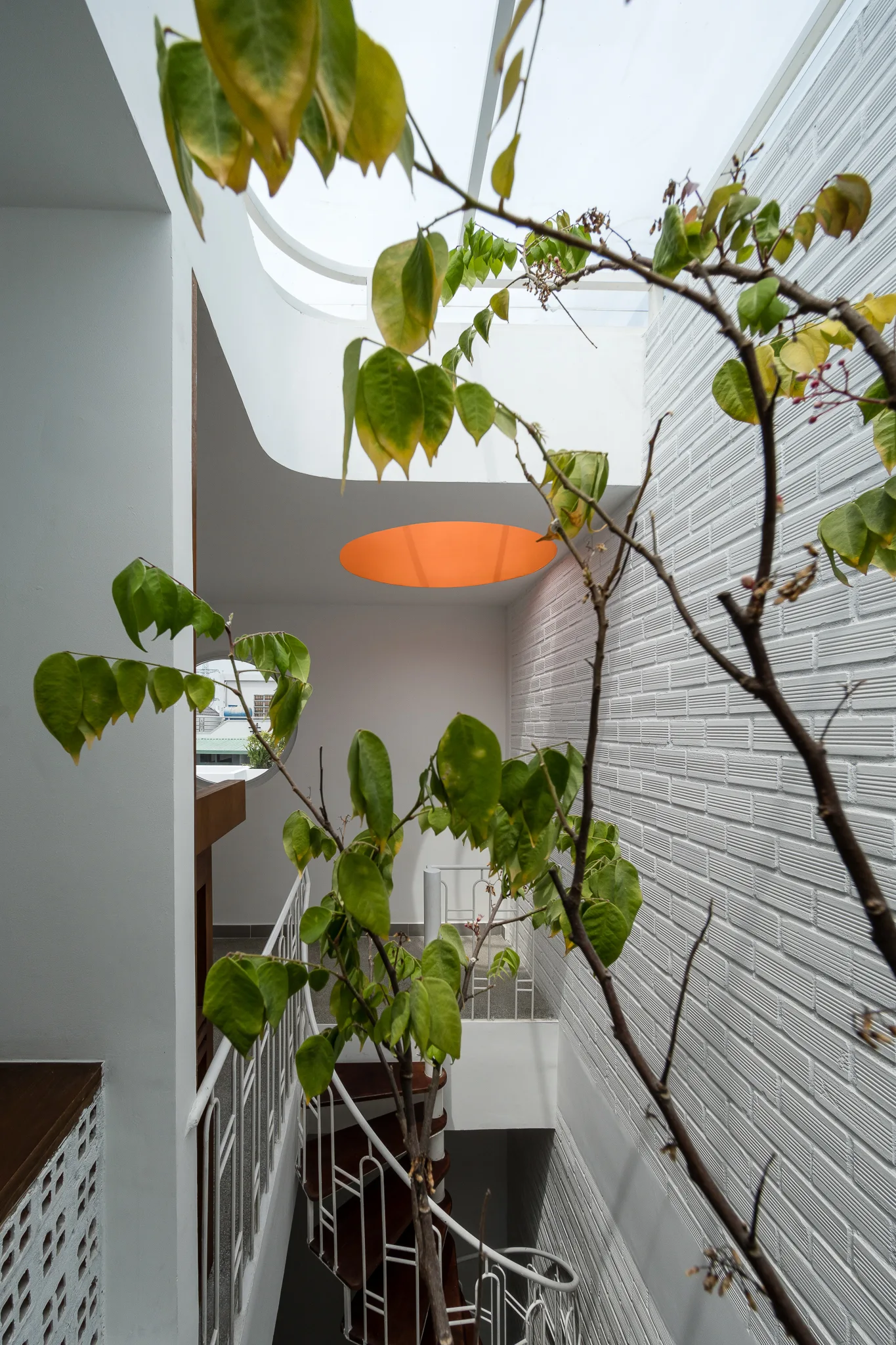 A spiral staircase with wooden treads and white railings stands against a white brick wall under a glass skylight with a circular orange accent.