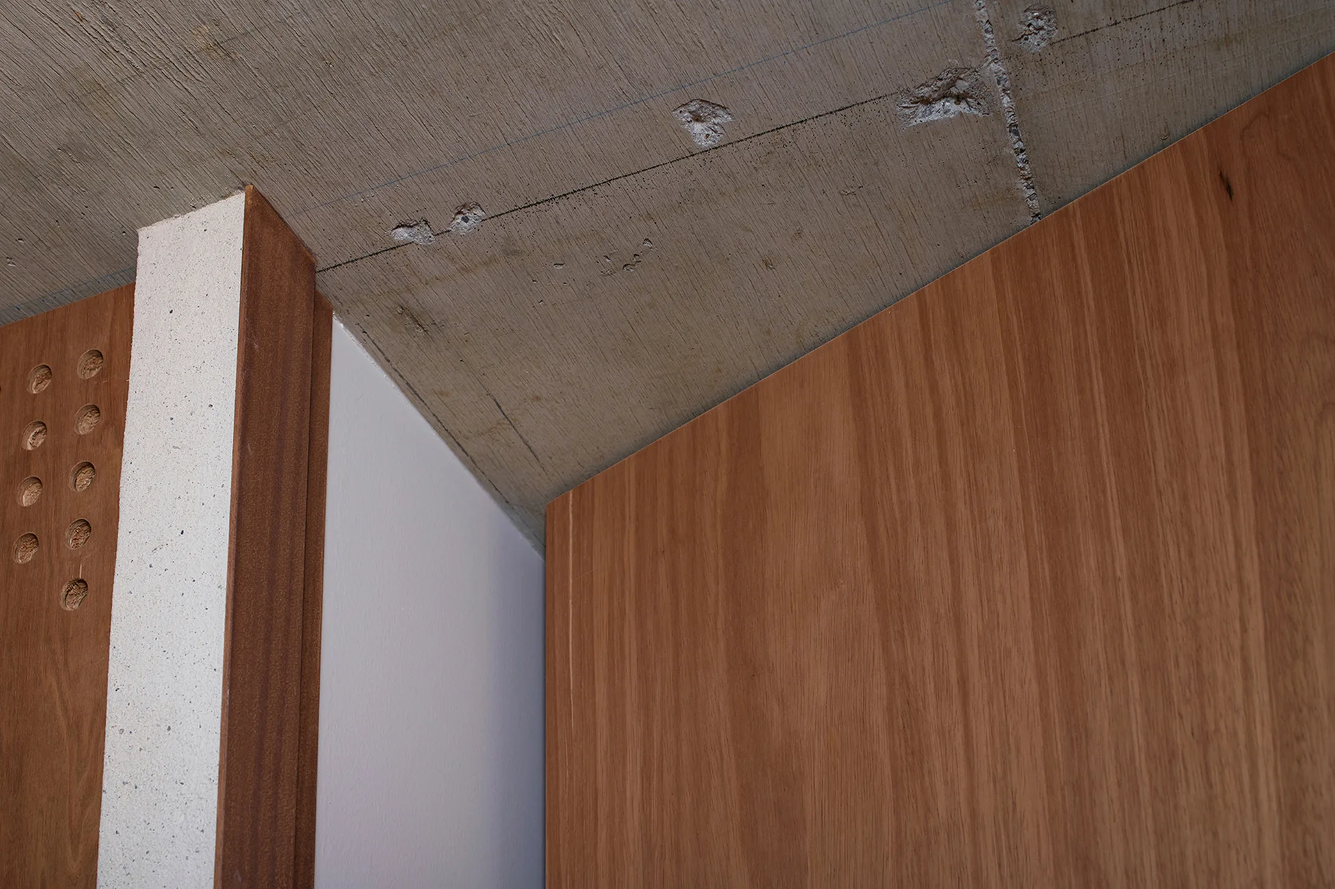 A close-up of vertical wood panels and a plaster partition wall meeting a raw, exposed concrete ceiling in an architectural corner.