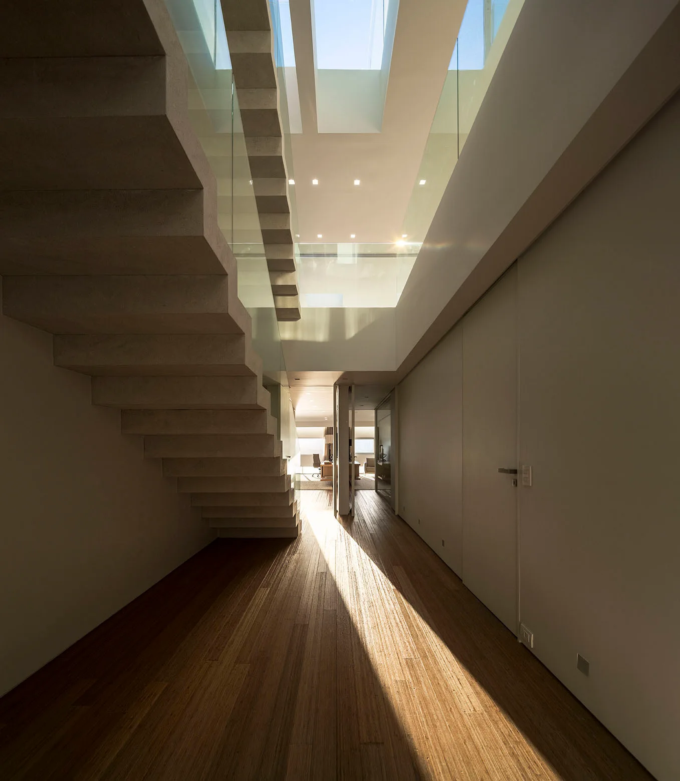A minimalist hallway features a floating concrete staircase with glass railings and wood flooring illuminated by overhead skylights.