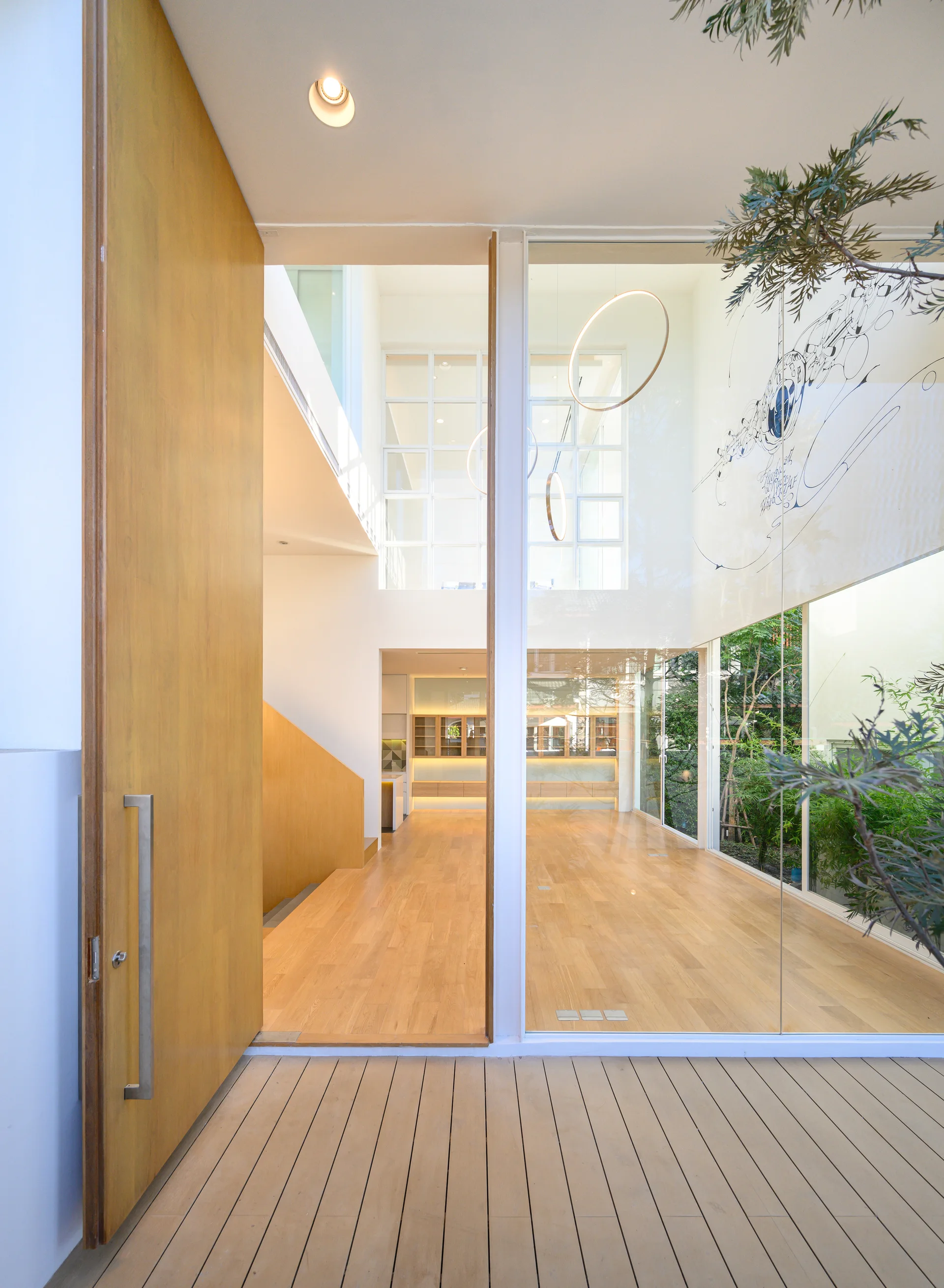A modern entry foyer features a large open wooden door, an outdoor wood deck, and a tall glass partition revealing wood floors and a staircase inside.