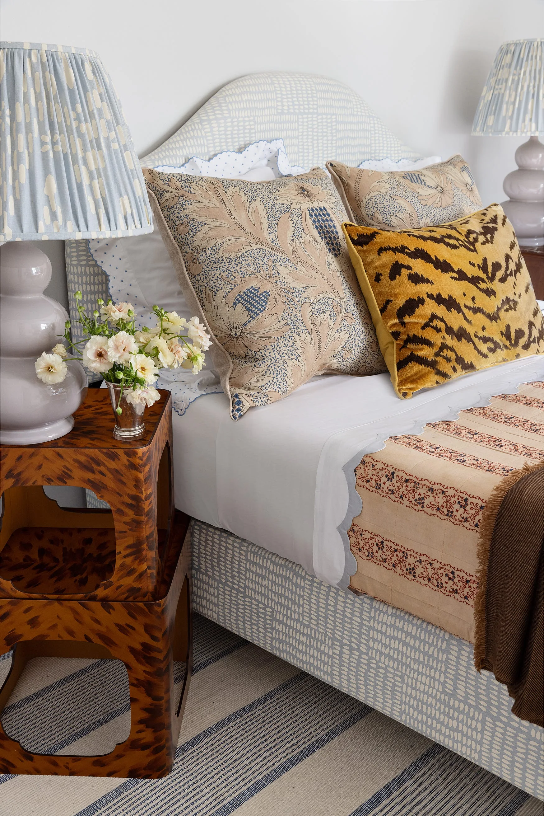 An upholstered blue and white headboard paired with a tortoiseshell bedside table, patterned throw pillows, and a ceramic lamp with a pleated shade.