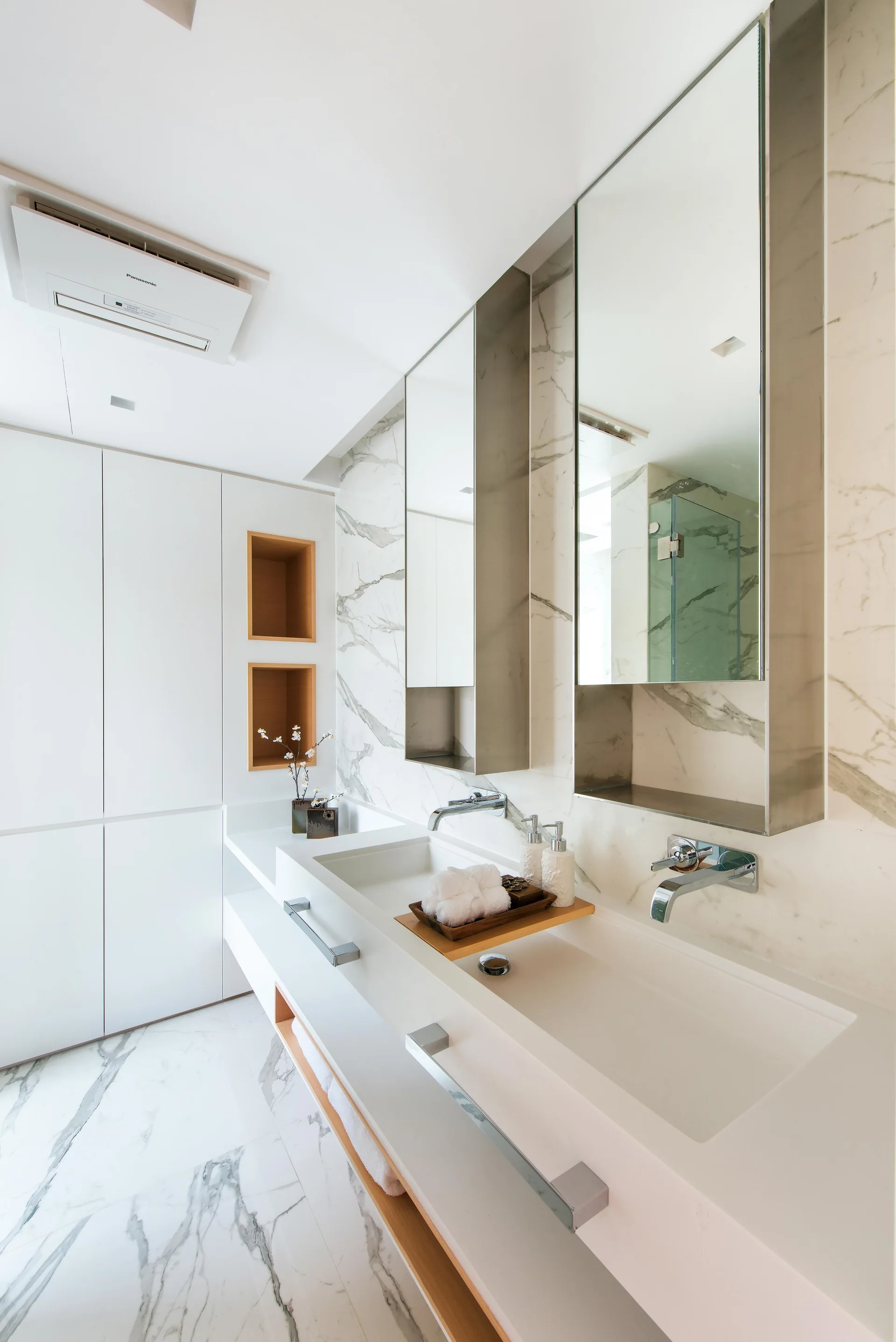 A modern bathroom features a white double sink vanity, wall-mounted silver faucets, marble tile walls, and recessed wood cubbies.