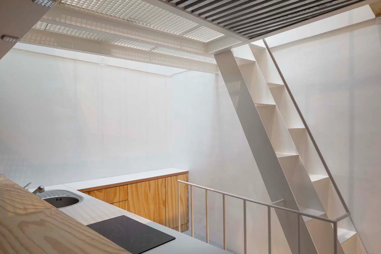 A minimalist loft kitchen features light plywood cabinets, a sink, and a white metal staircase rising toward a slatted overhead skylight.