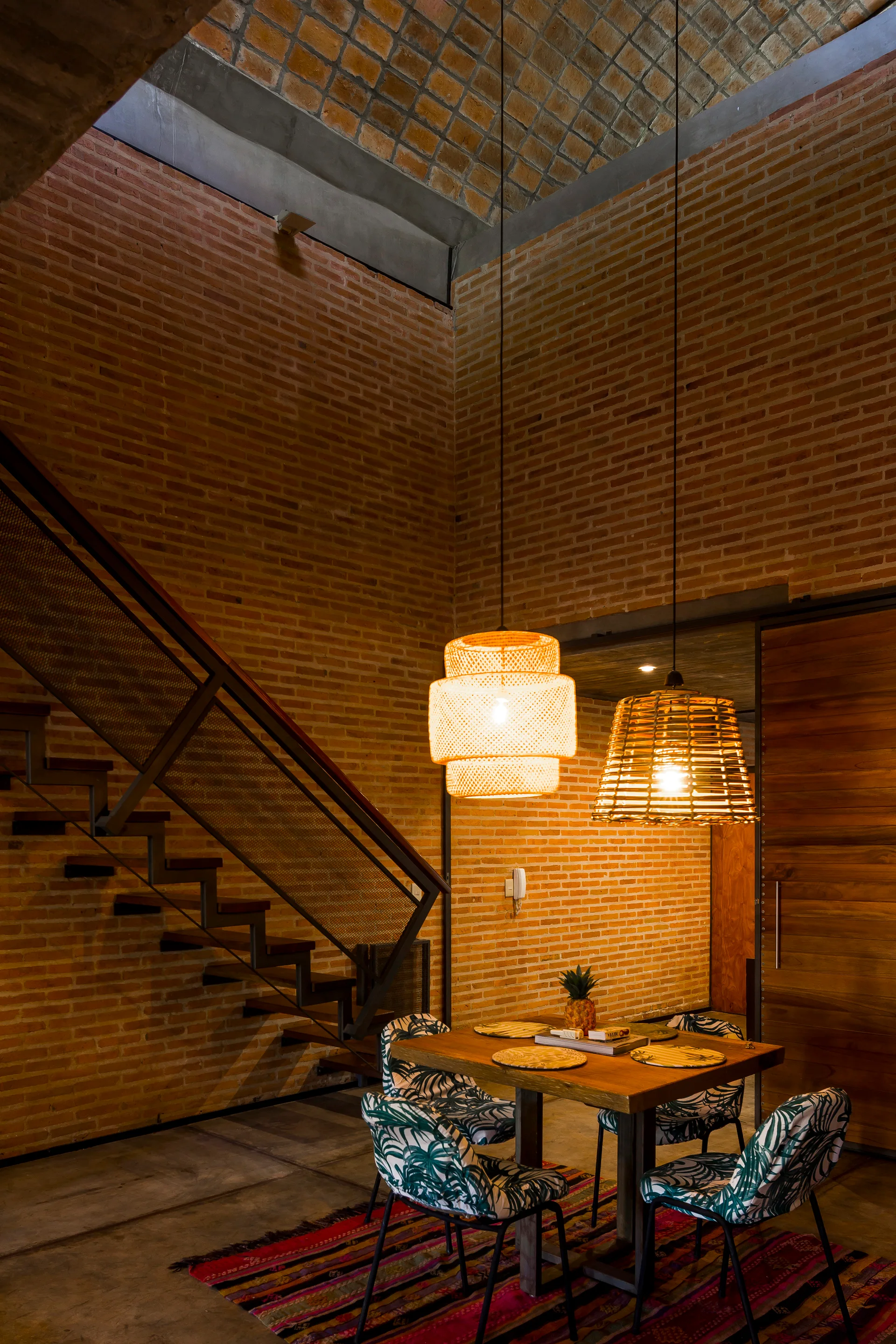 A dining table with tropical print chairs sits under two woven pendant lights in a room featuring high exposed brick walls and a metal staircase.