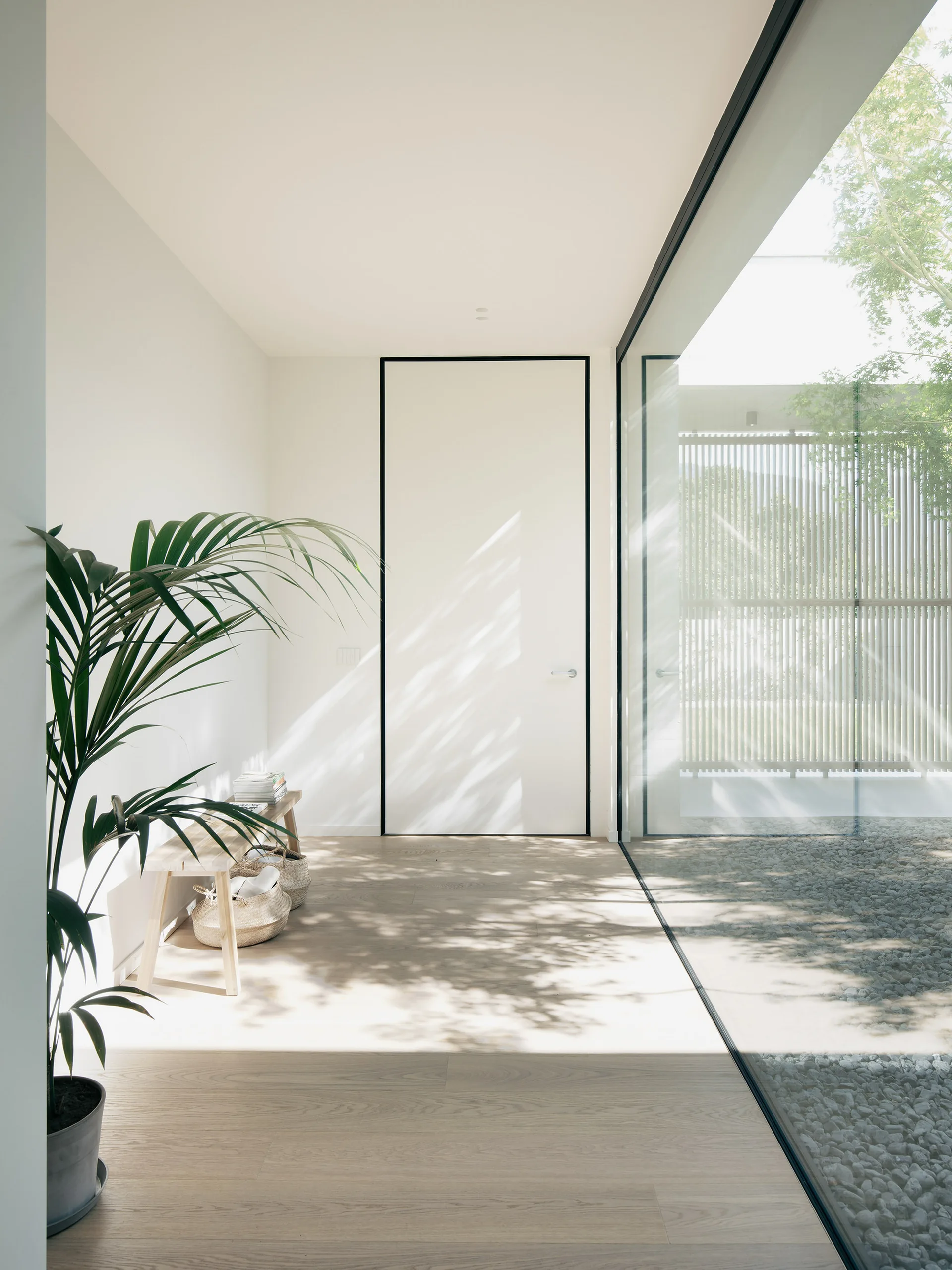 A minimalist entryway features a light wood floor, a wooden bench with baskets, a tall black-framed door, and a large glass wall.