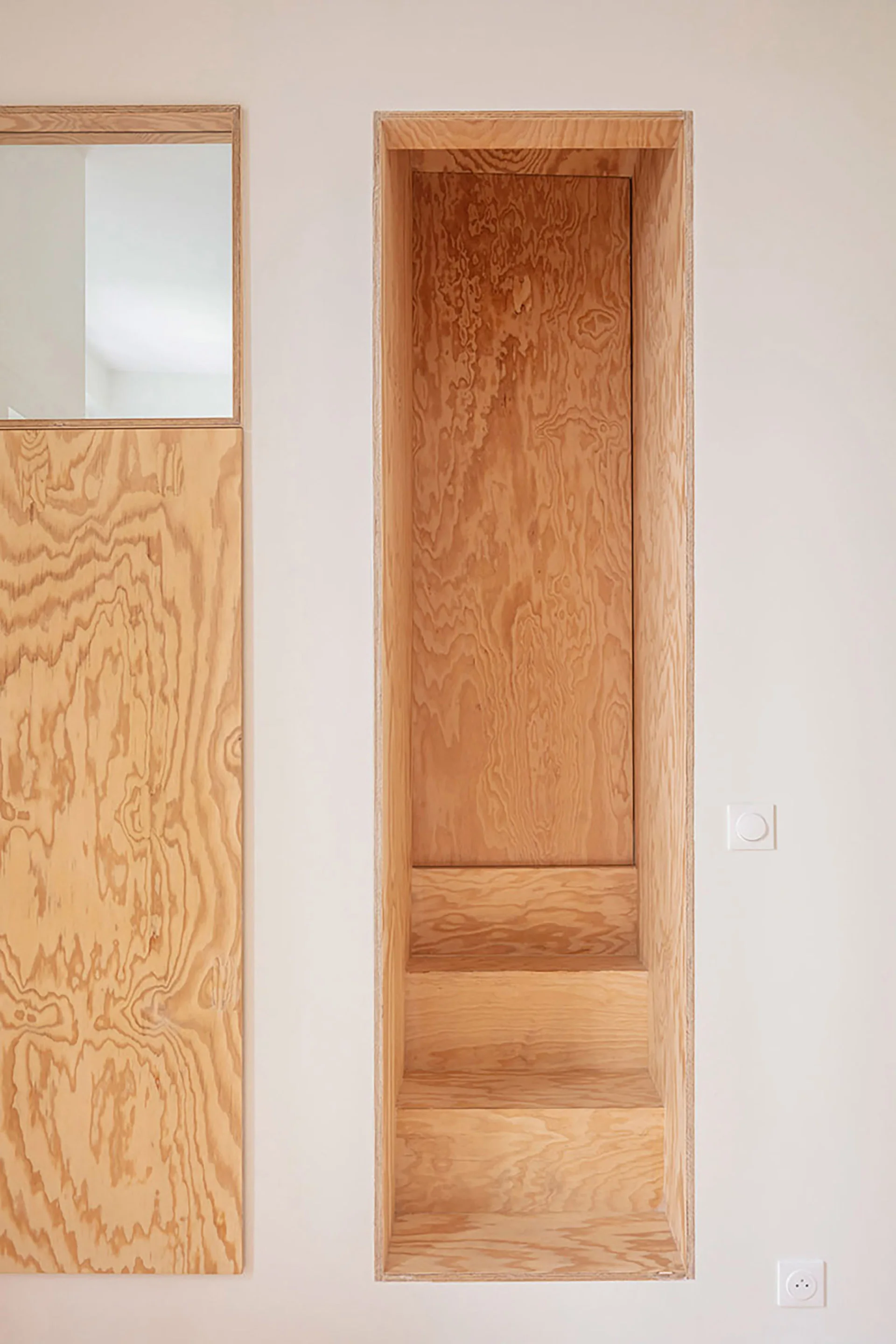 A recessed staircase crafted from light-toned plywood is built into a smooth white wall next to a matching wooden wall panel.