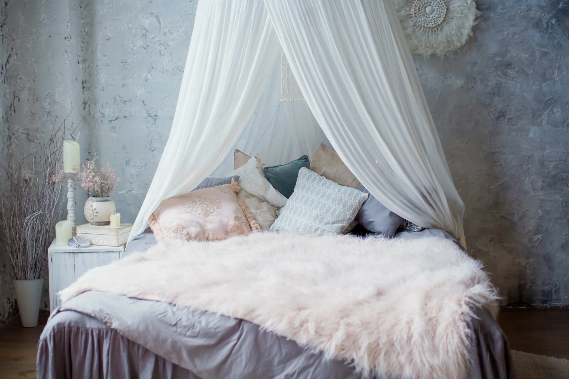 A bed with a sheer white canopy, layered pastel pillows, and a pink faux fur throw stands against a textured gray plaster wall.