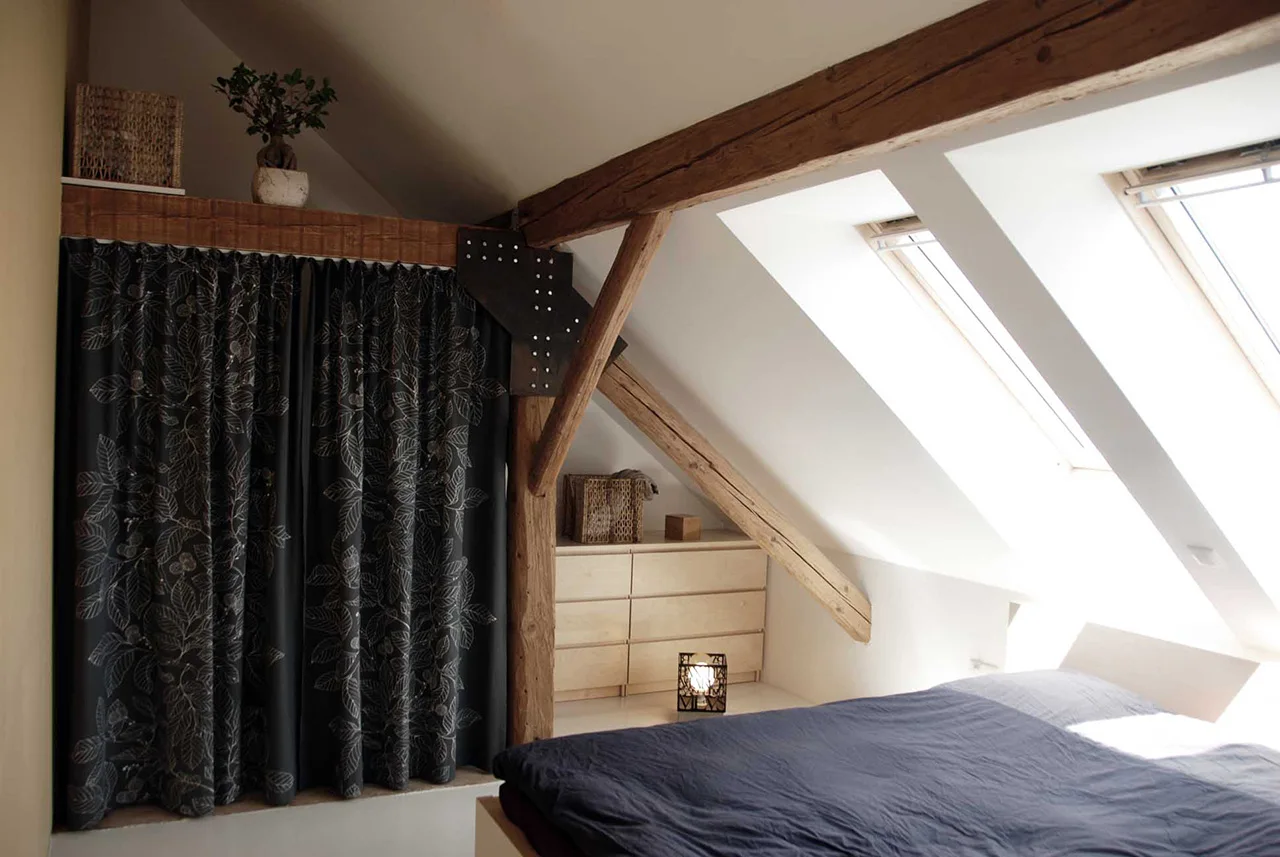 A bright attic bedroom with sloped ceilings and skylights, featuring exposed rustic wooden beams, a low bed, and dark patterned curtains.