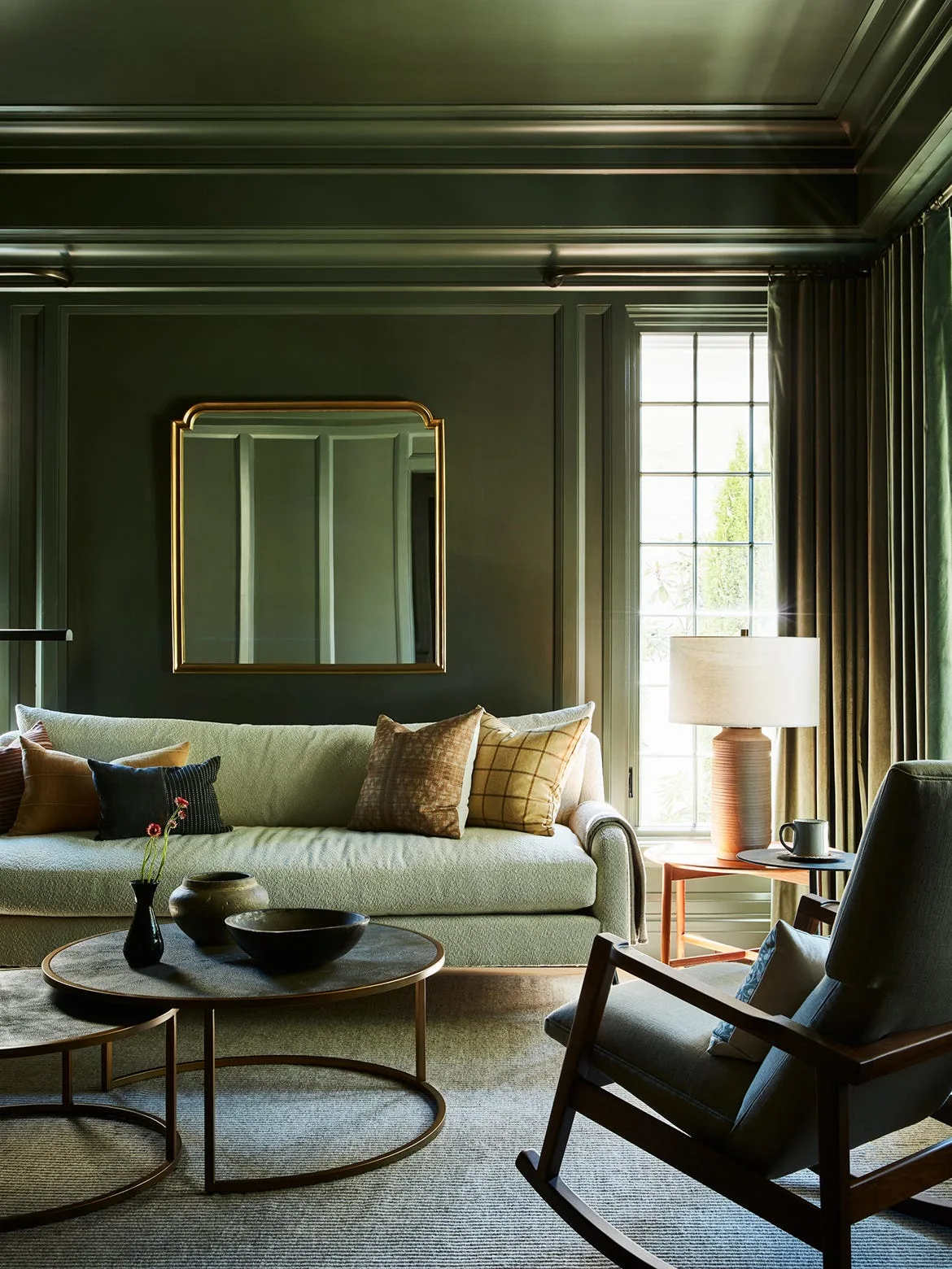 A moody living room with paneled dark green walls and ceiling, a light green upholstered sofa, round nested coffee tables, and a gold-framed mirror.