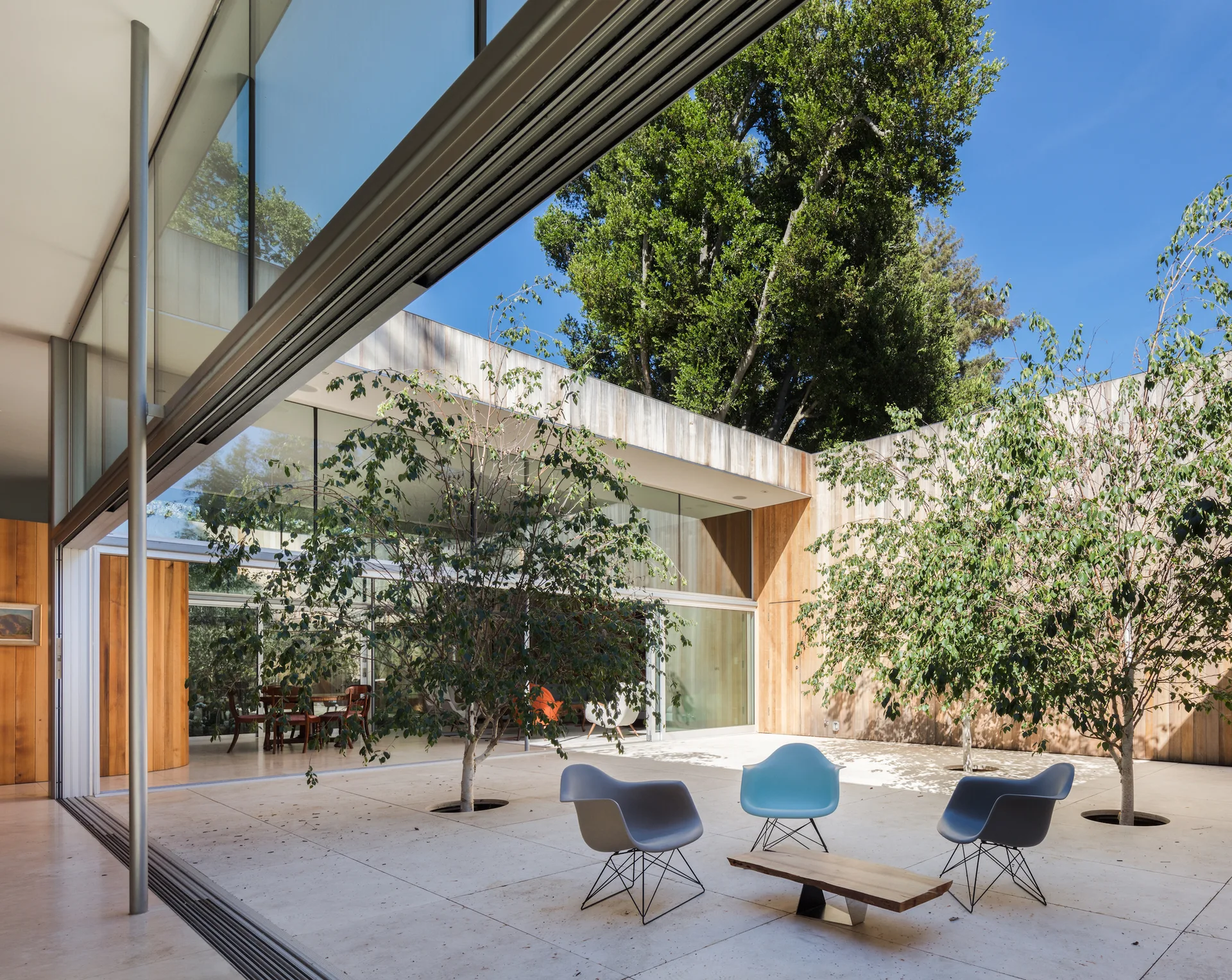 A modern open-air courtyard features molded plastic chairs, a low wood coffee table, and live trees integrated into the beige stone tile floor.
