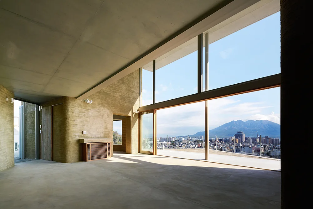 A minimalist room with rough tan concrete walls, a smooth grey ceiling, and a massive glass window wall framing a mountain and city view.