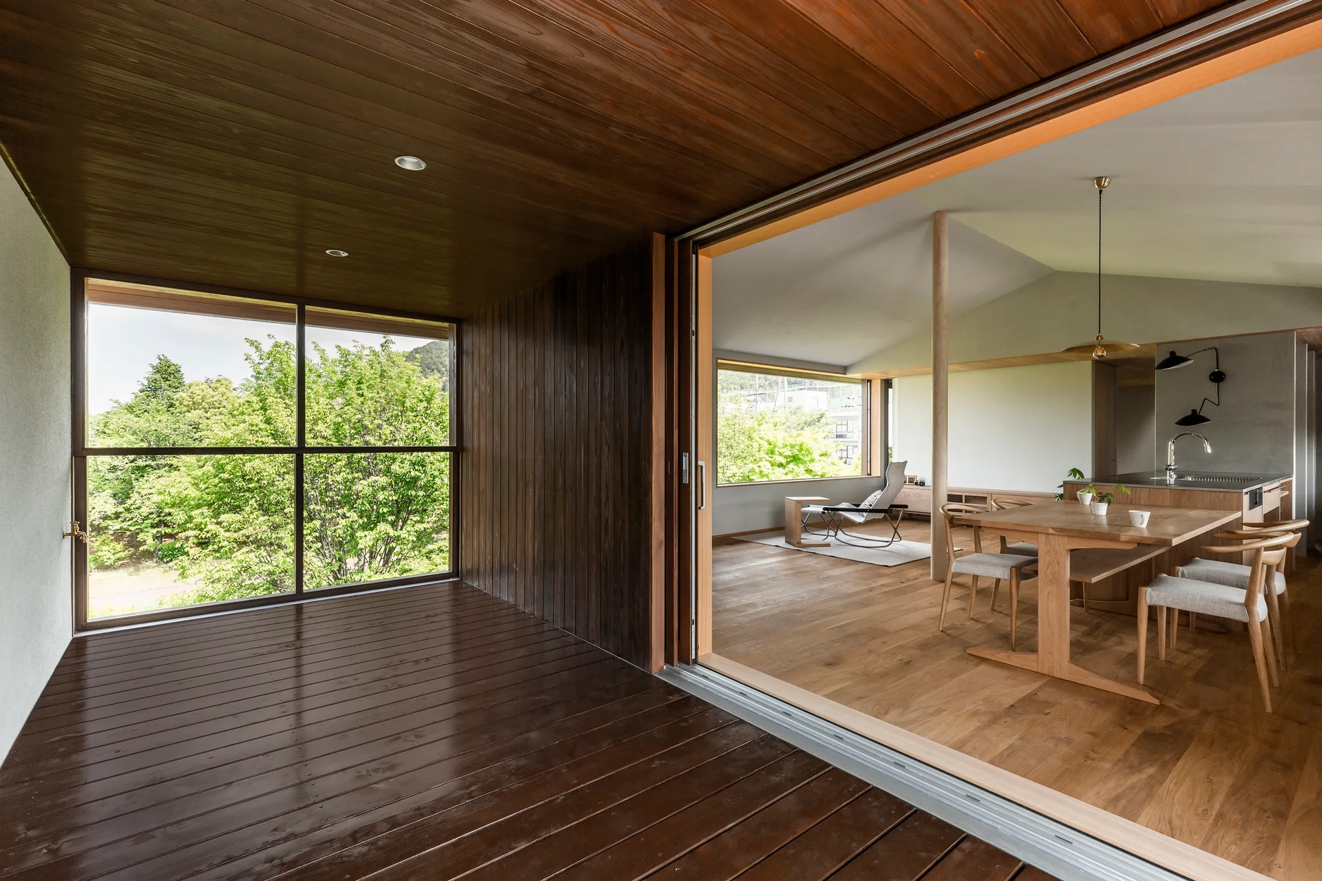 A dark wood covered deck looks through open sliding glass doors into a bright dining room with a light oak table and chairs.