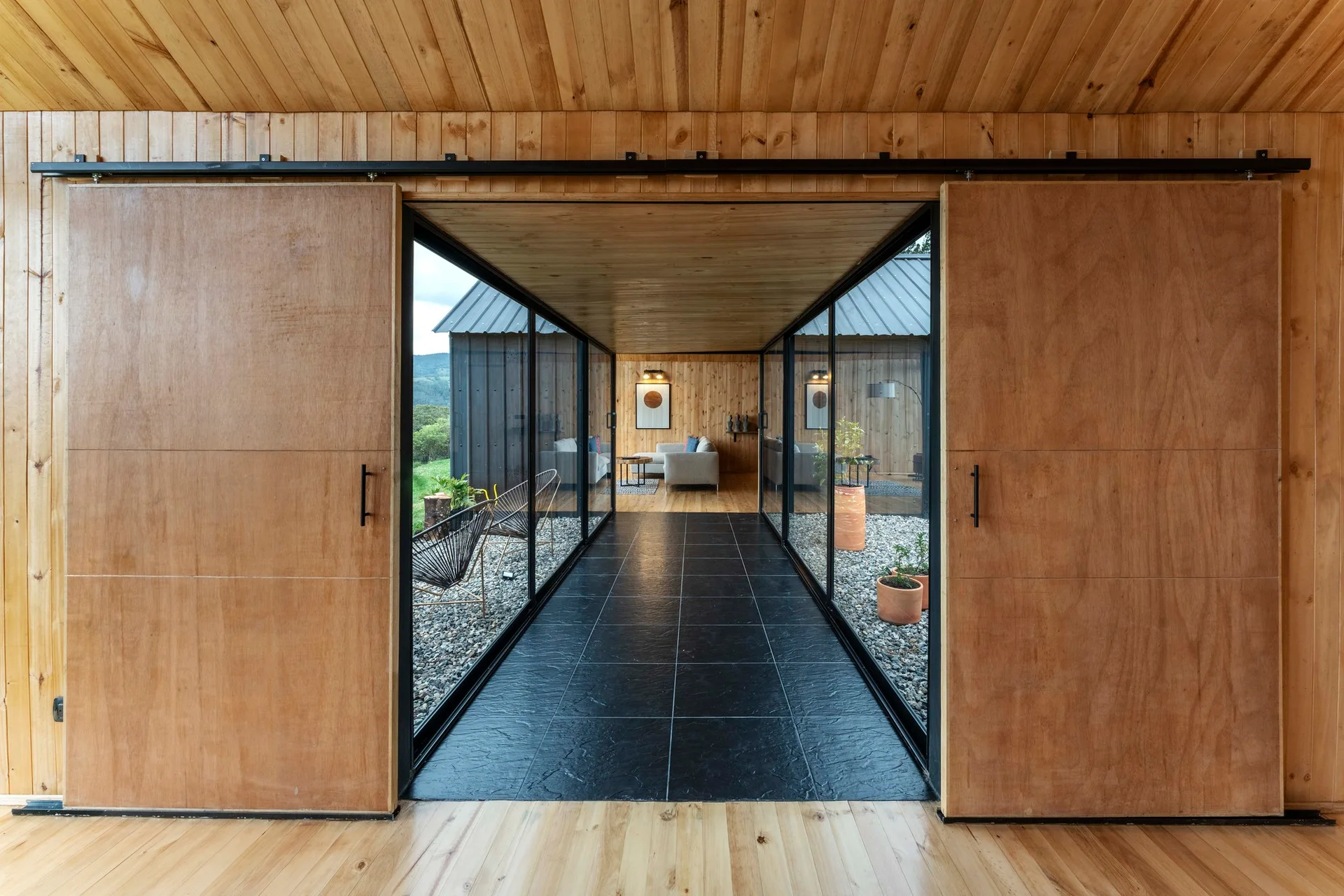 A modern transition hallway featuring glass walls overlooking gravel courtyards framed by wooden sliding doors and dark stone floor tiles.