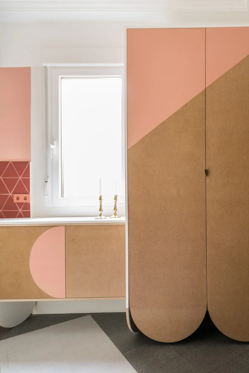 Modern kitchen cabinetry featuring custom doors with pink and natural wood geometric shapes, set beside a white window and patterned pink wall tiles.