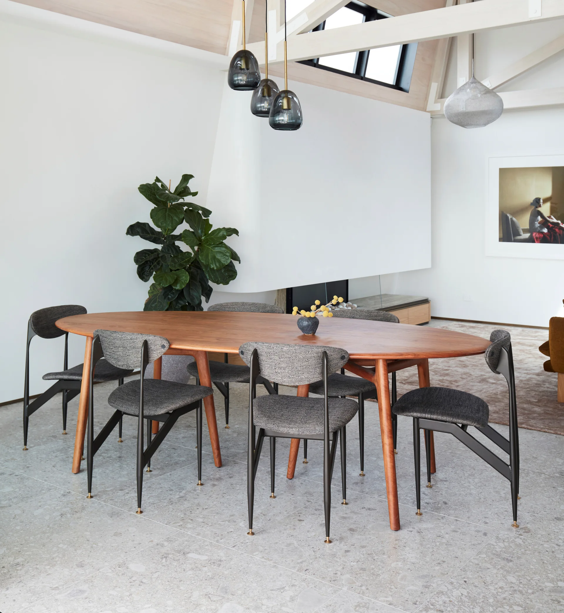 A modern dining area features an oval wood table with upholstered grey chairs on a speckled stone tile floor under white ceiling beams.
