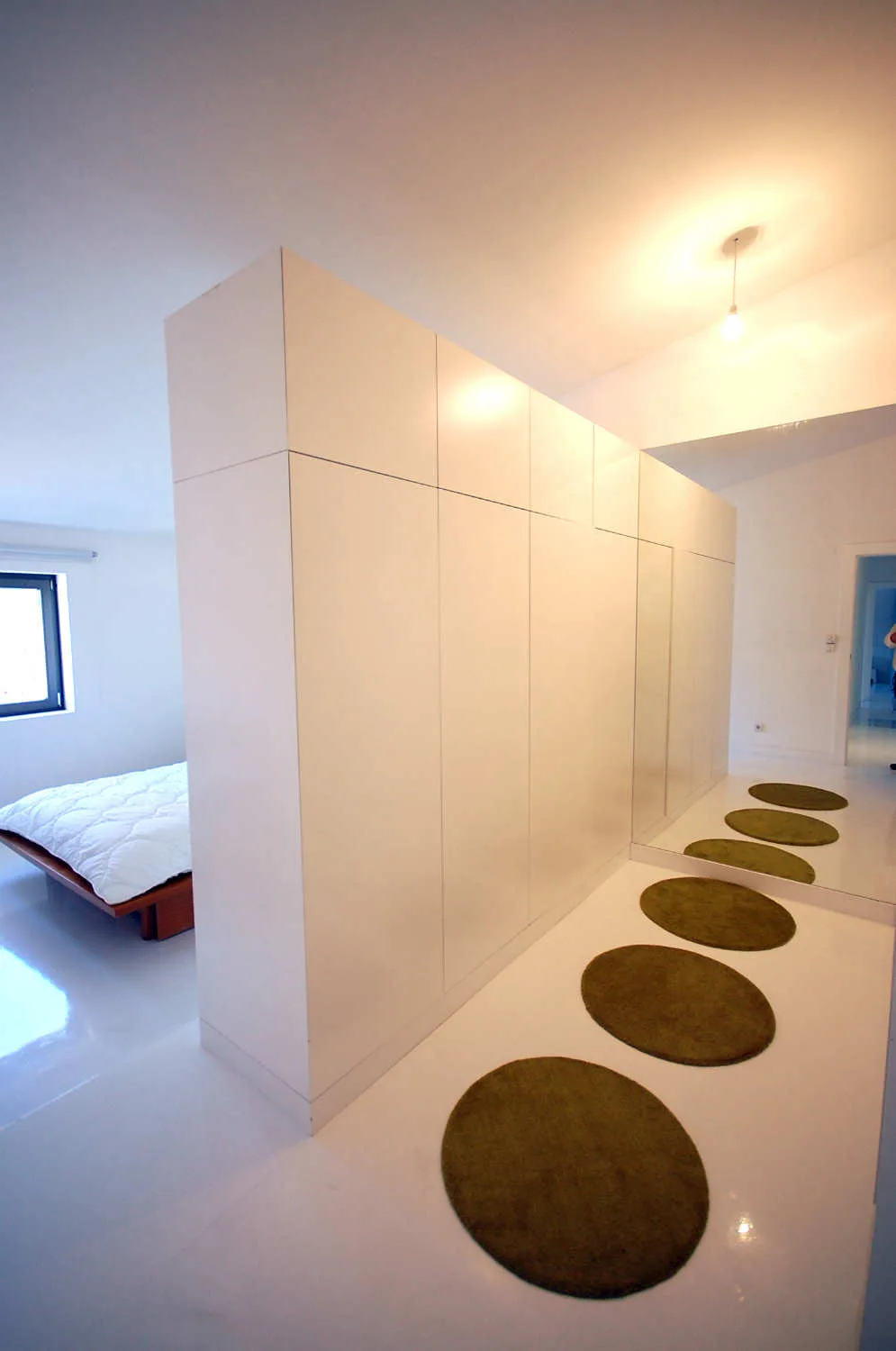 A minimalist bedroom features a tall white wardrobe acting as a room divider, with a row of circular green rugs on a glossy white floor.