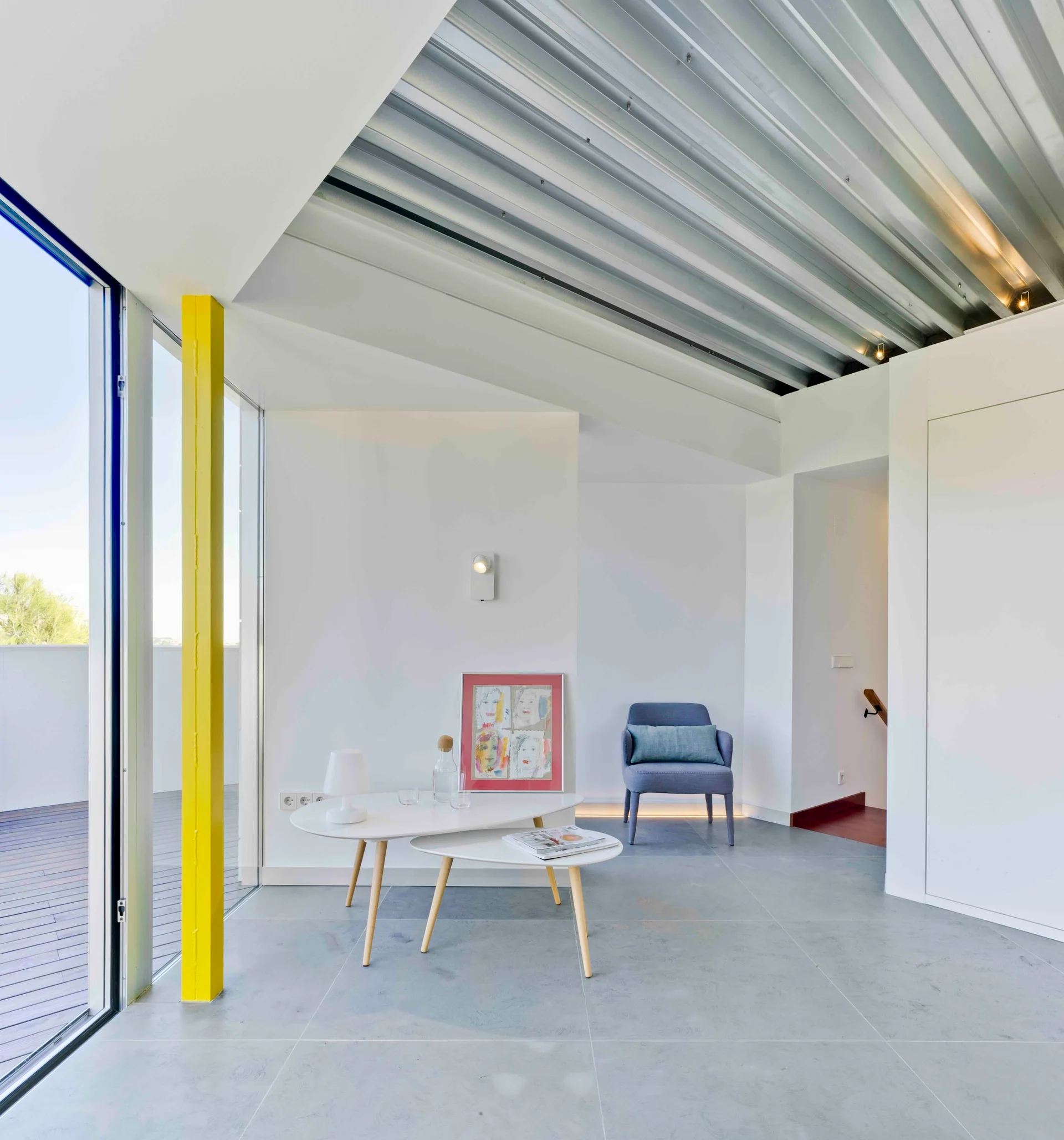 A modern white living room featuring a bright yellow column, a corrugated metal ceiling, nested coffee tables, and a blue armchair.