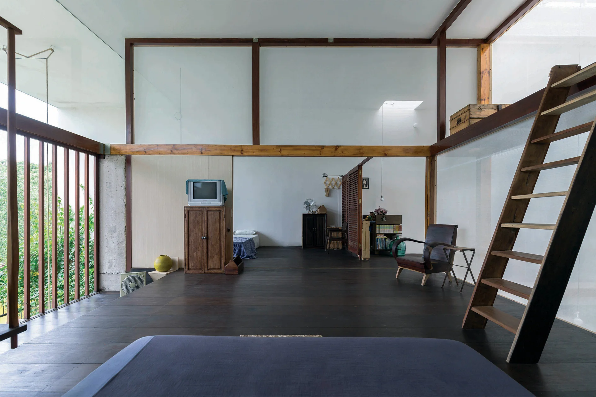 A minimalist mezzanine bedroom featuring dark wood flooring, a wooden structural post and beam frame, and a ladder.