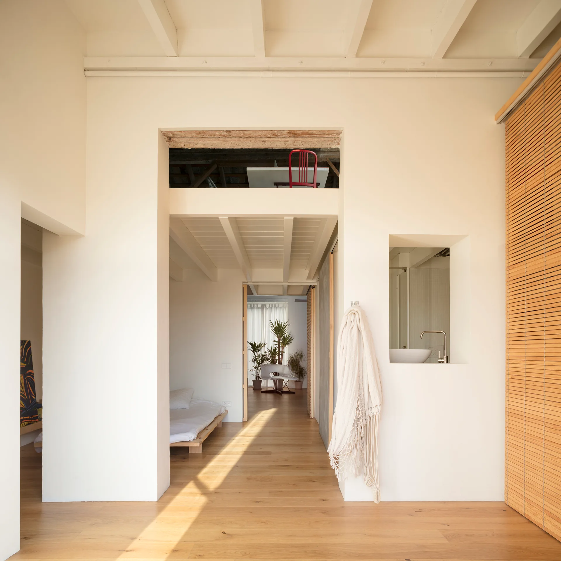An open-plan minimalist apartment features light wood floors, white plaster walls with exposed ceiling beams, a built-in sink niche, and a daybed.